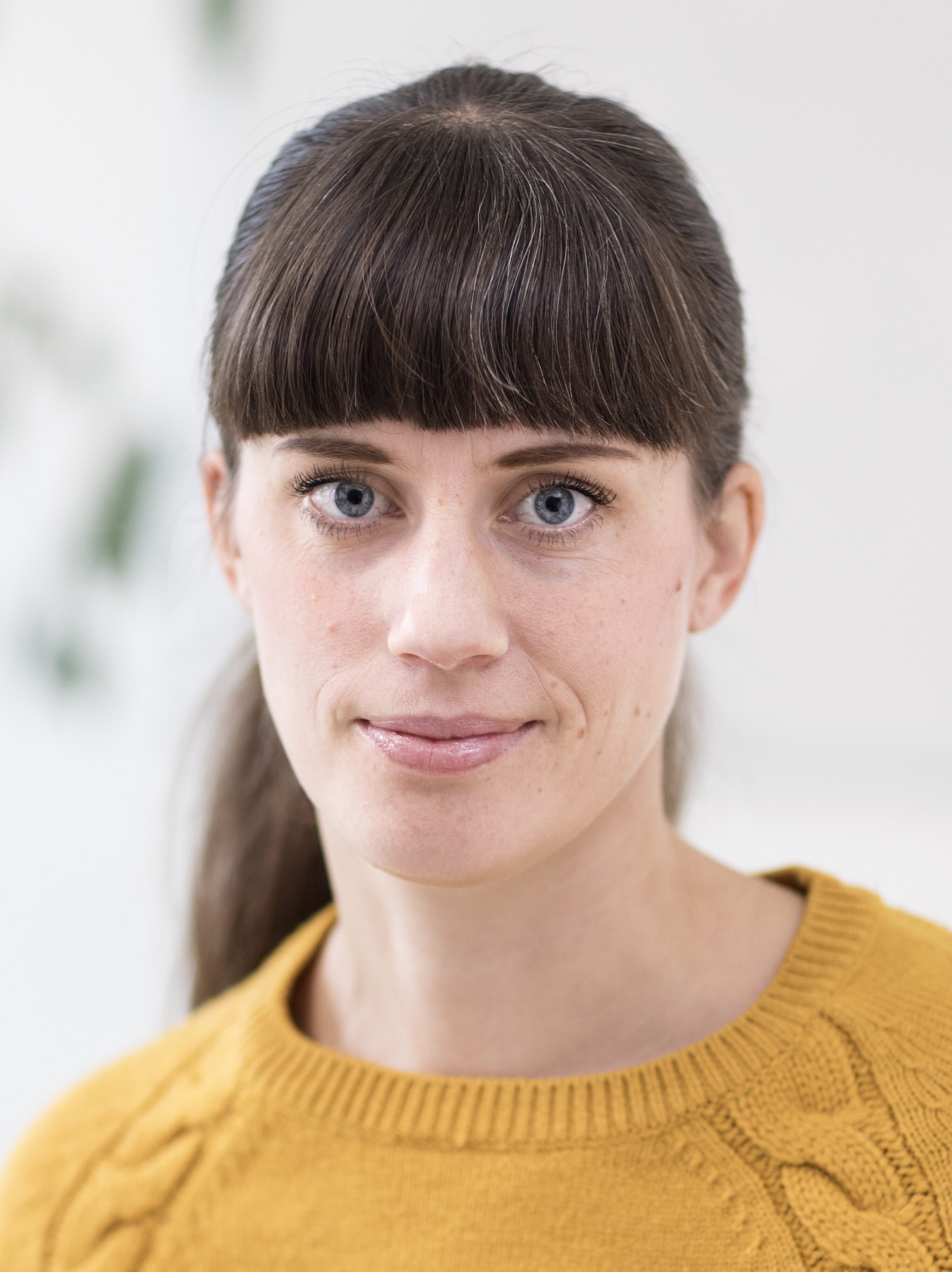 En kvinna med rakt brunt hår och lugg, har en gul tröja på sig och tittar in i kameran med neutral min. Bakgrunden är suddig.