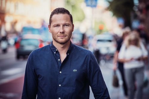 Fredrik Norberg i en blå skjorta står på en stadsgata med människor och trafik i bakgrunden.