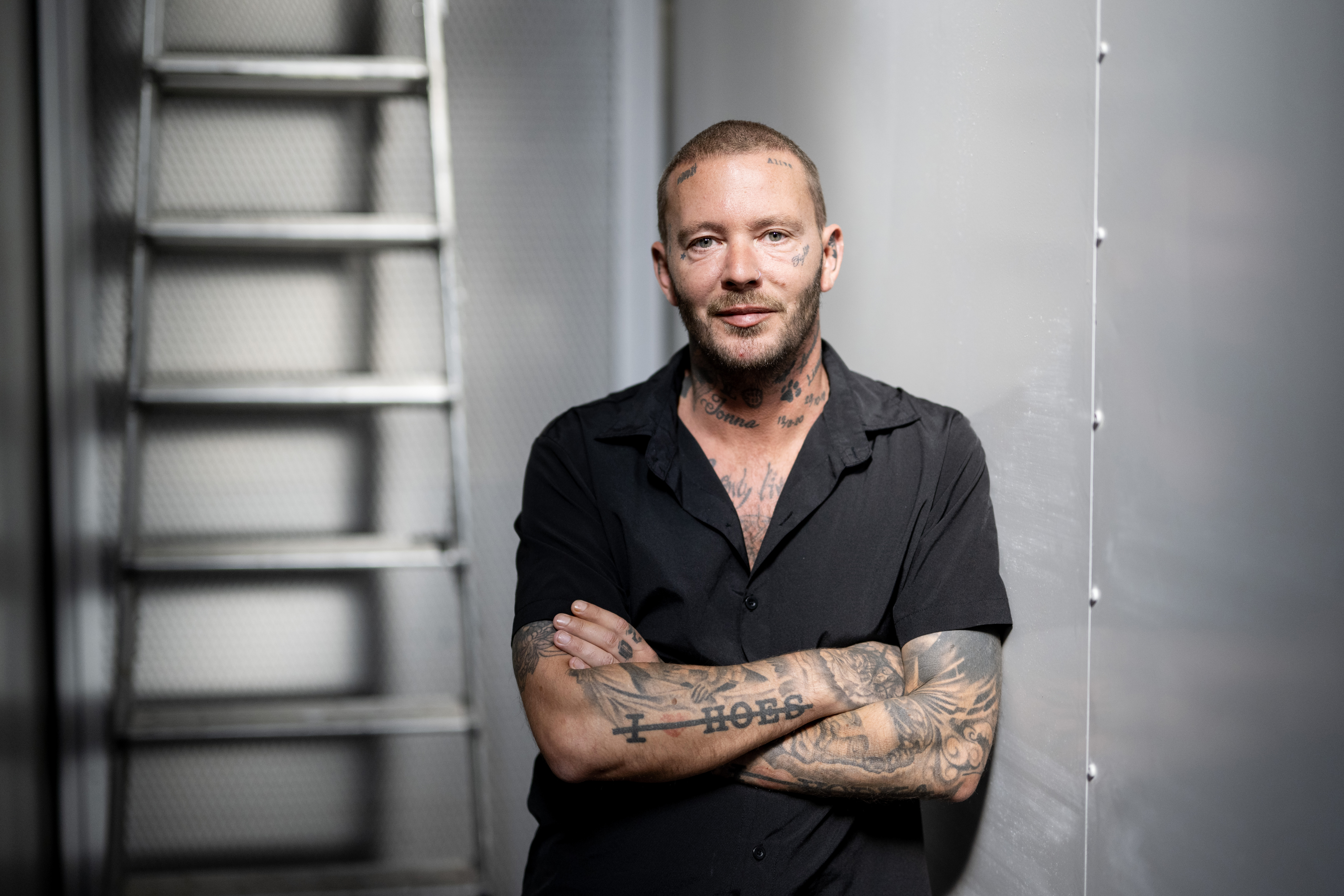 Man med rakat huvud och tatueringar i svart skjorta tittar i kameran.