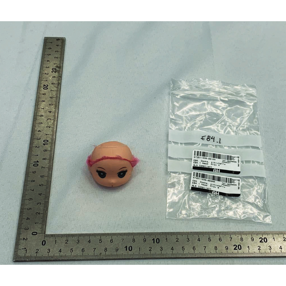 Ett dockhuvud med rosa hår ligger bredvid en märkt plastpåse och två metallinjal som bildar ett L på en vit yta.