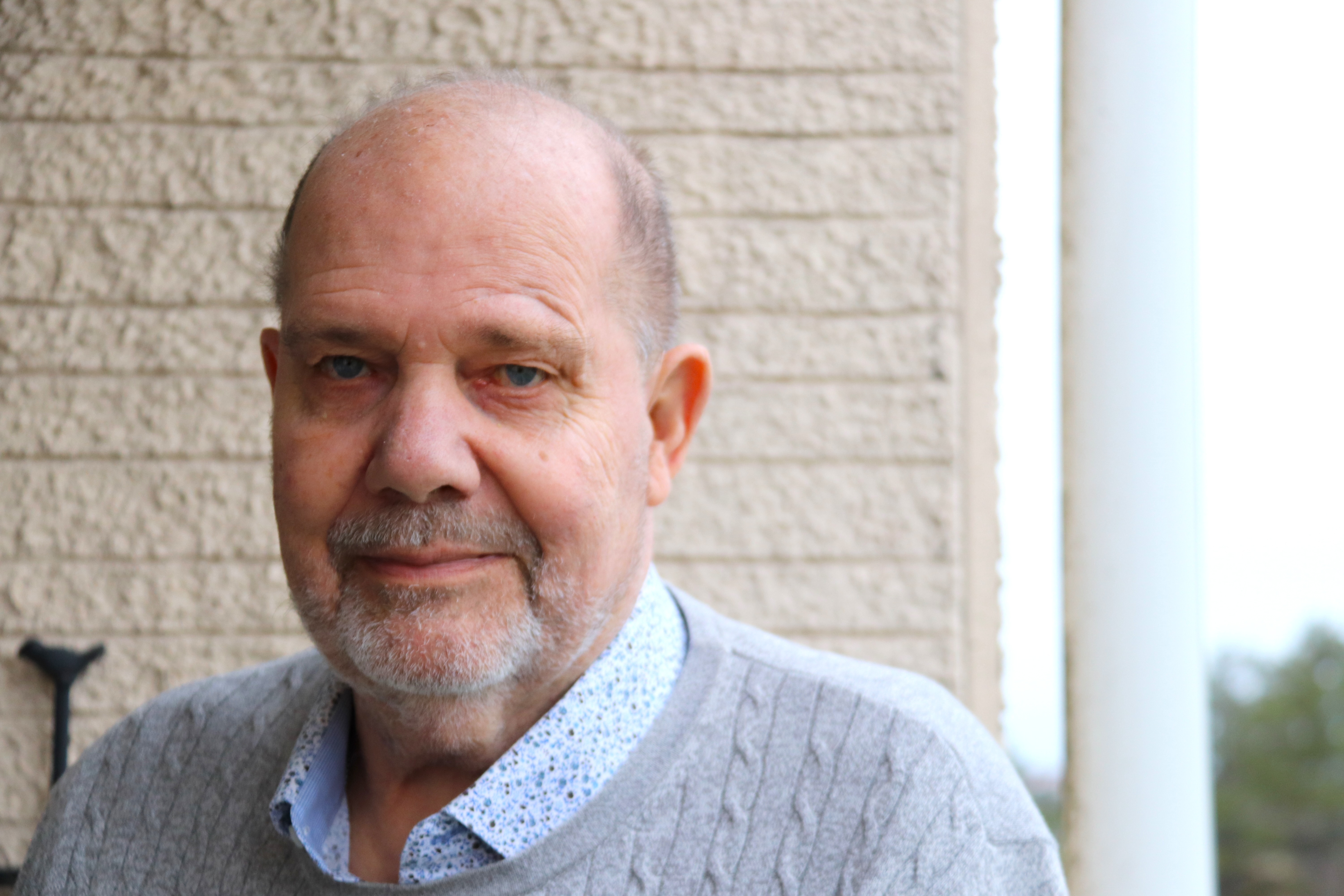 En äldre man med skalligt huvud och grått skägg har en grå tröja och mönstrad skjorta, står framför en beige vägg.