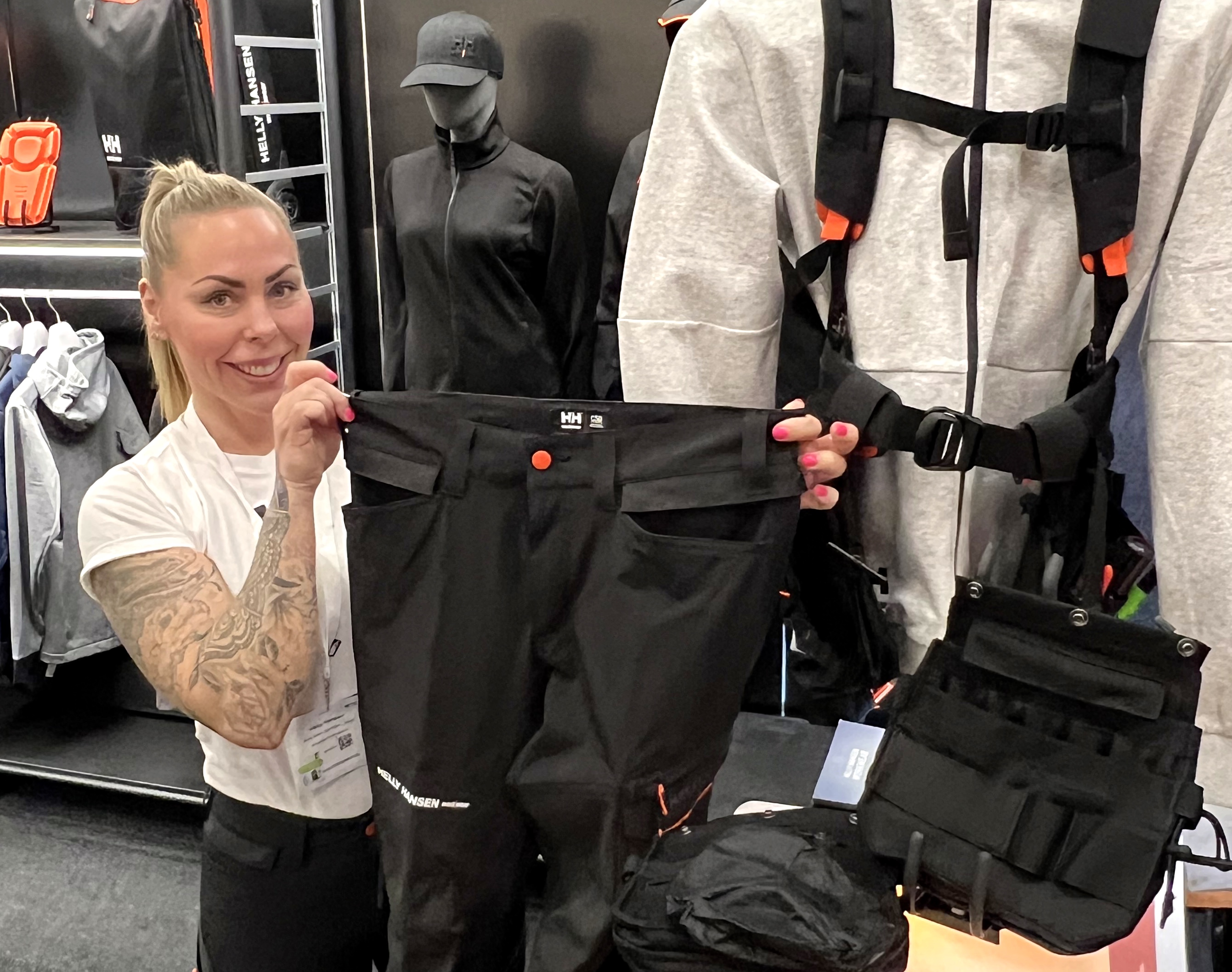En tatuerad kvinna håller upp ett par svarta Helly Hansen-byxor vid en monter för arbetskläder och tillbehör.