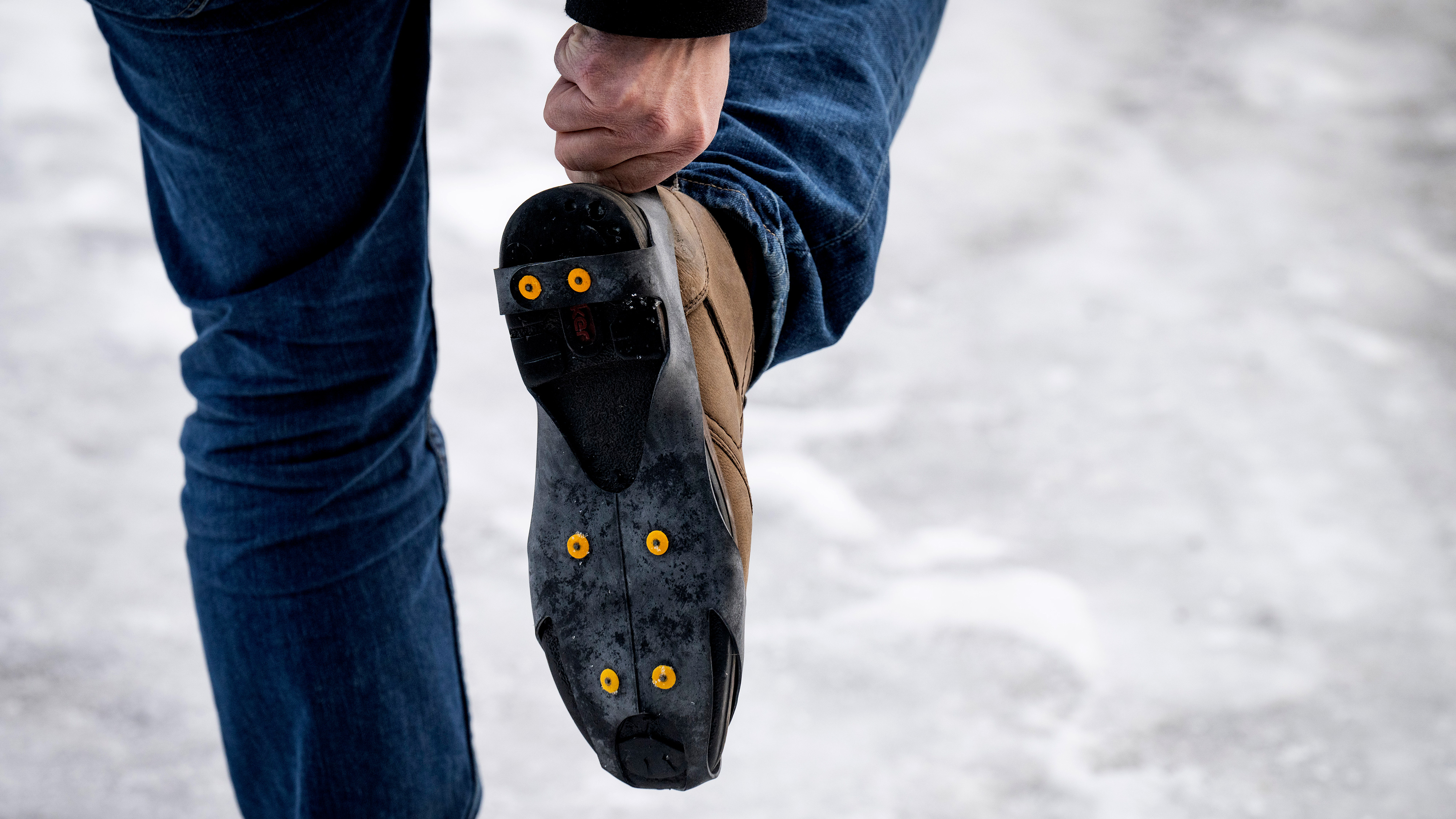 En person med jeans håller upp en stövel och visar halkskydd med gula dubbar som sitter fast på sulan för att gå på is.