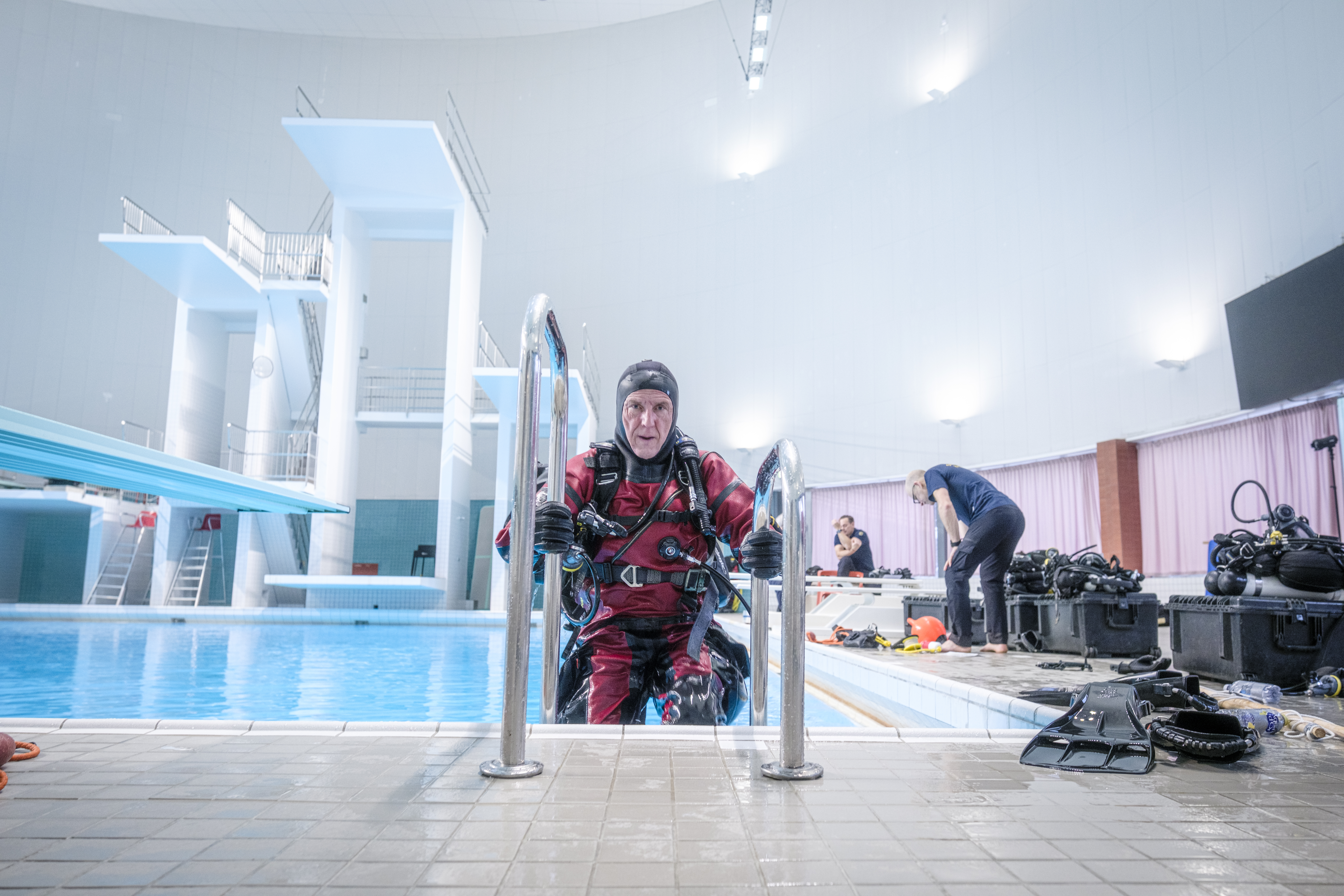 En person i röd och svart våtdräkt klättrar upp ur en inomhuspool medan en annan ordnar dykutrustning intill.