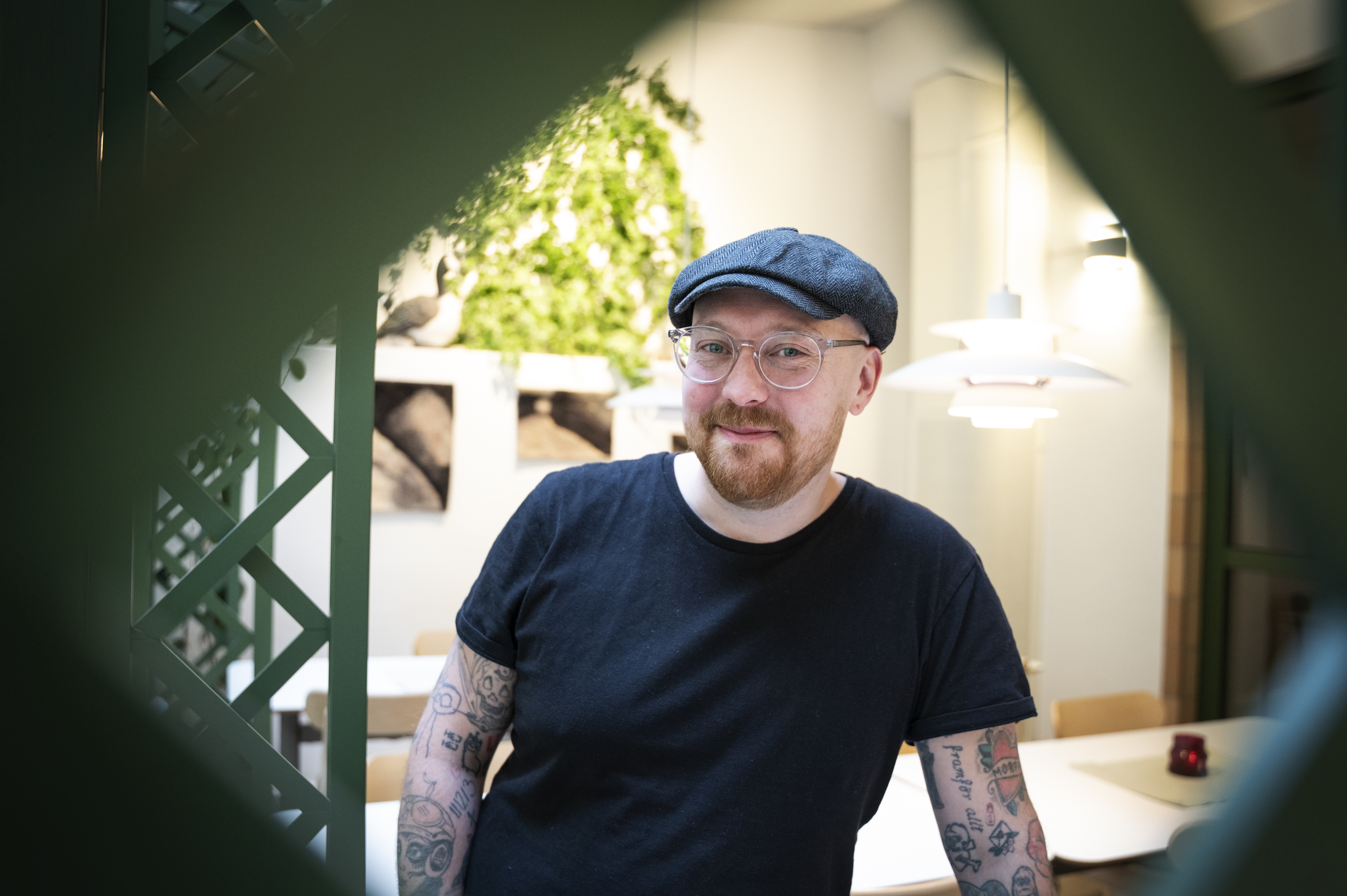En man med glasögon, kepsar och tatueringar på armarna står inne. Bakom honom finns växter och inramade foton.