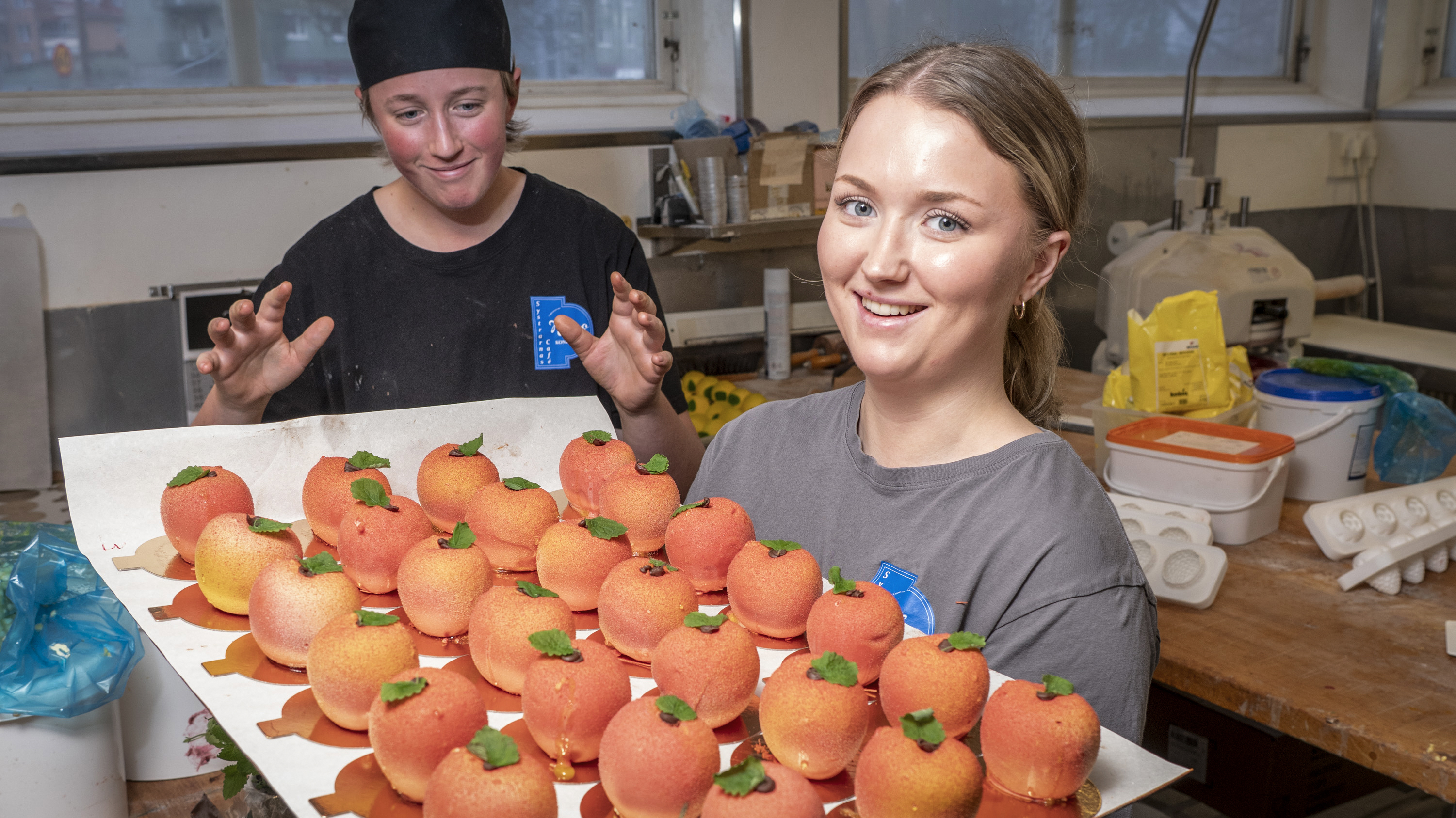 Två personer i ett kök, en håller en bricka med runda, orange bakverk med gröna stjälkar. Kökstillbehör och ingredienser syns i bakgrunden.