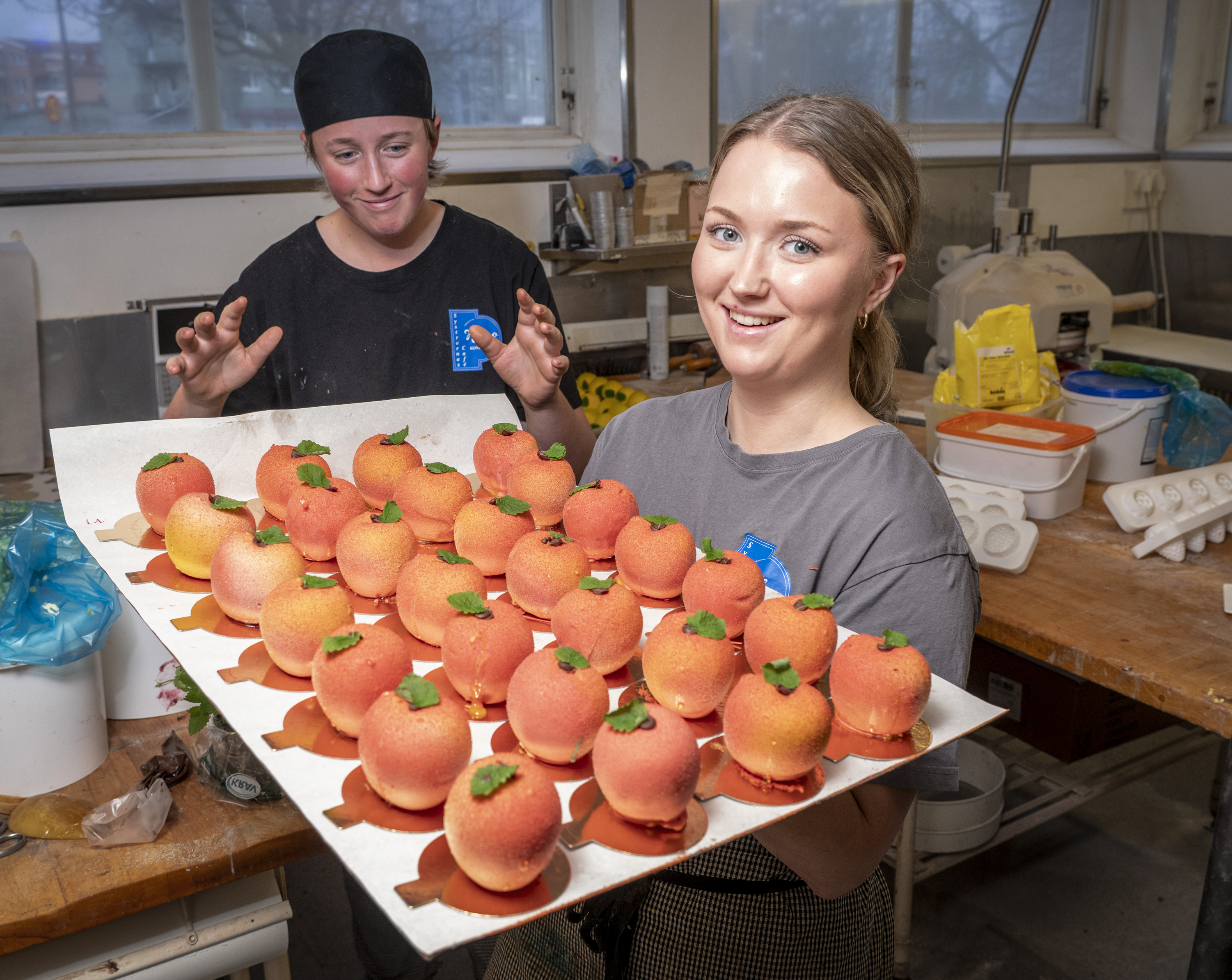Två personer i ett kök, en håller en bricka med runda, orange bakverk med gröna stjälkar. Kökstillbehör och ingredienser syns i bakgrunden.