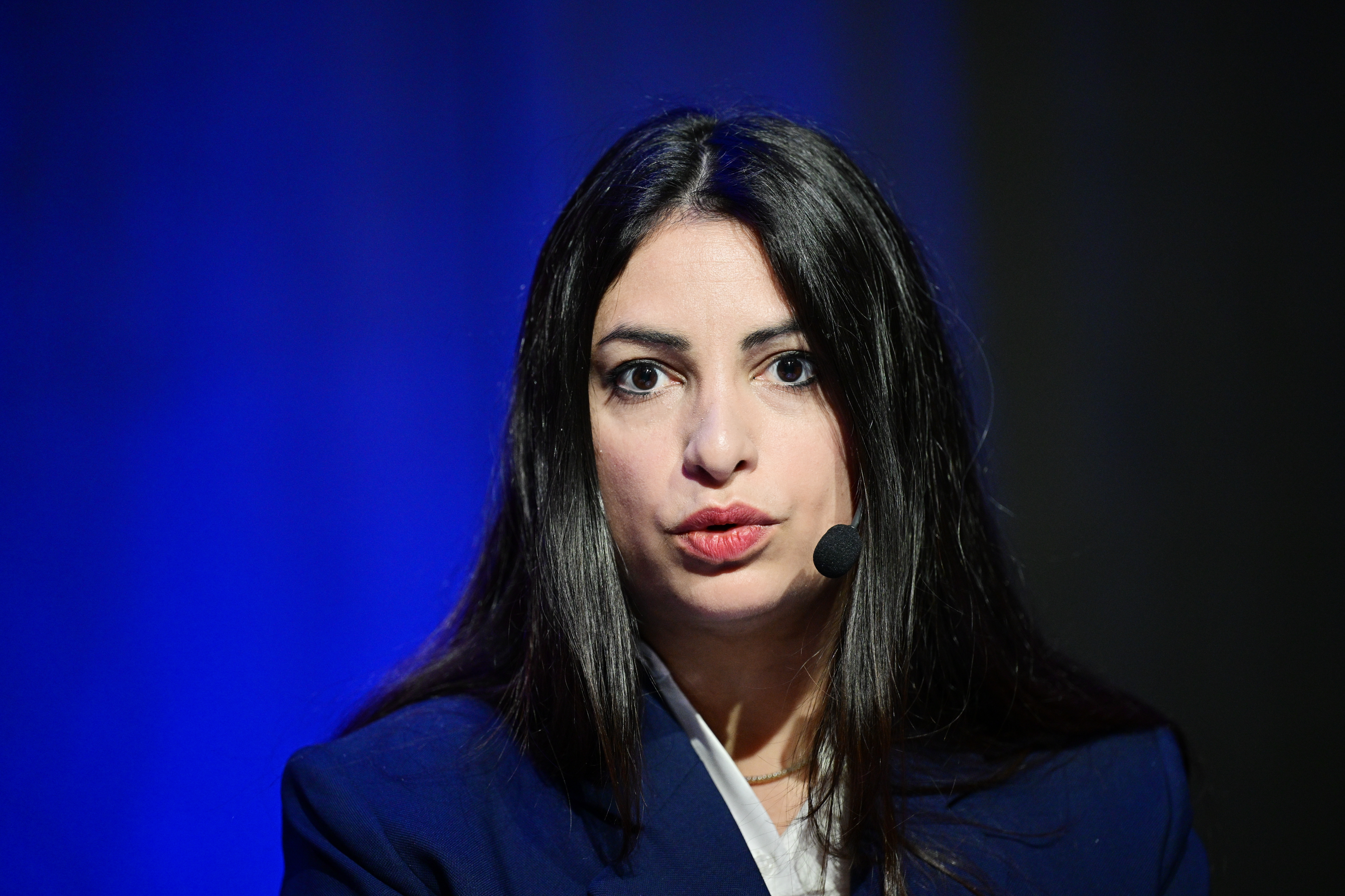 En kvinna med långt mörkt hår, klädd i en mörkblå kavaj och headsetmikrofon, talar framför en blå bakgrund.