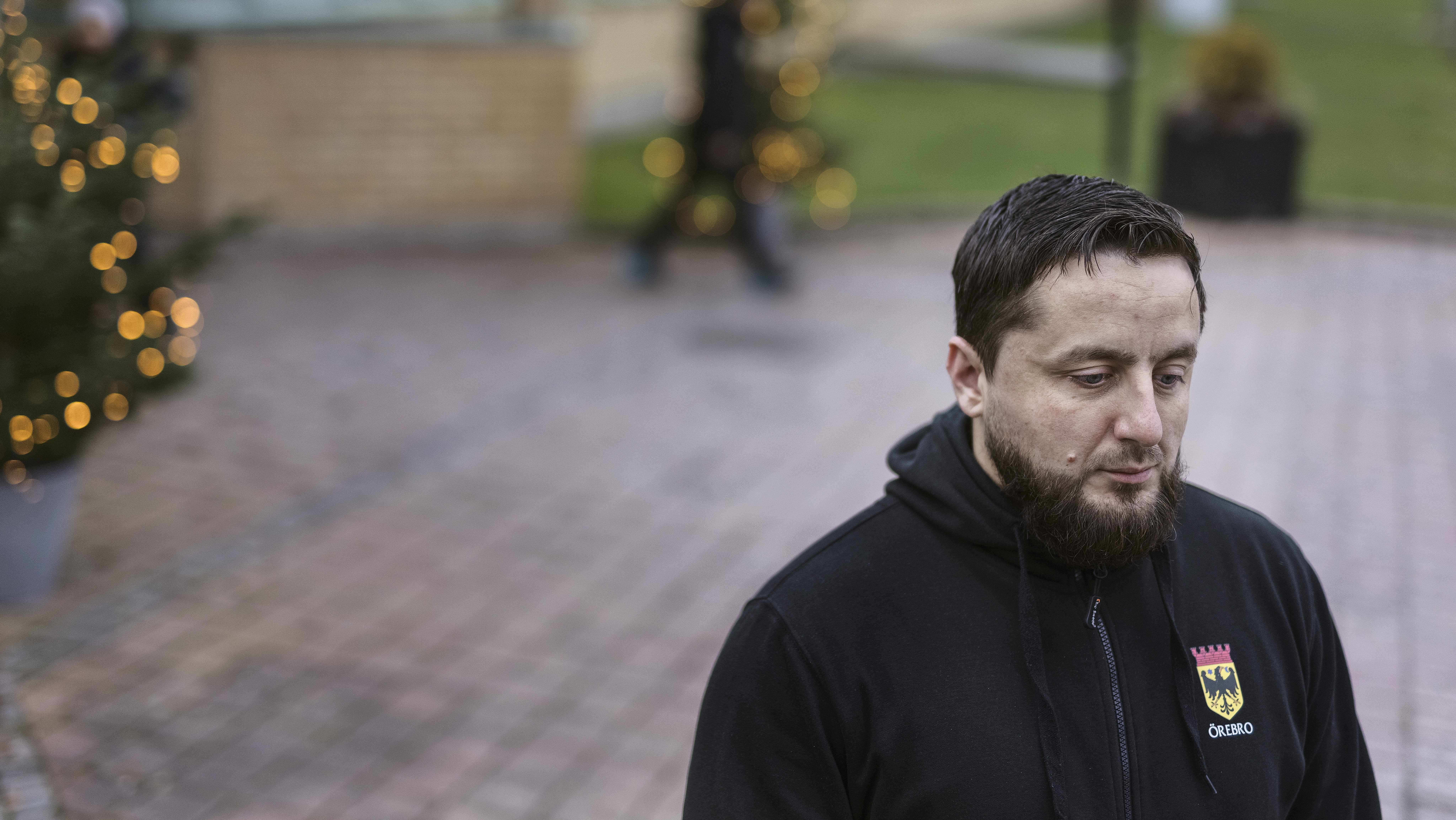 Adnan Imsirovic med skägg, bär en svart jacka med ett märke, står utomhus på en gångväg av tegel nära julgranar med ljus.