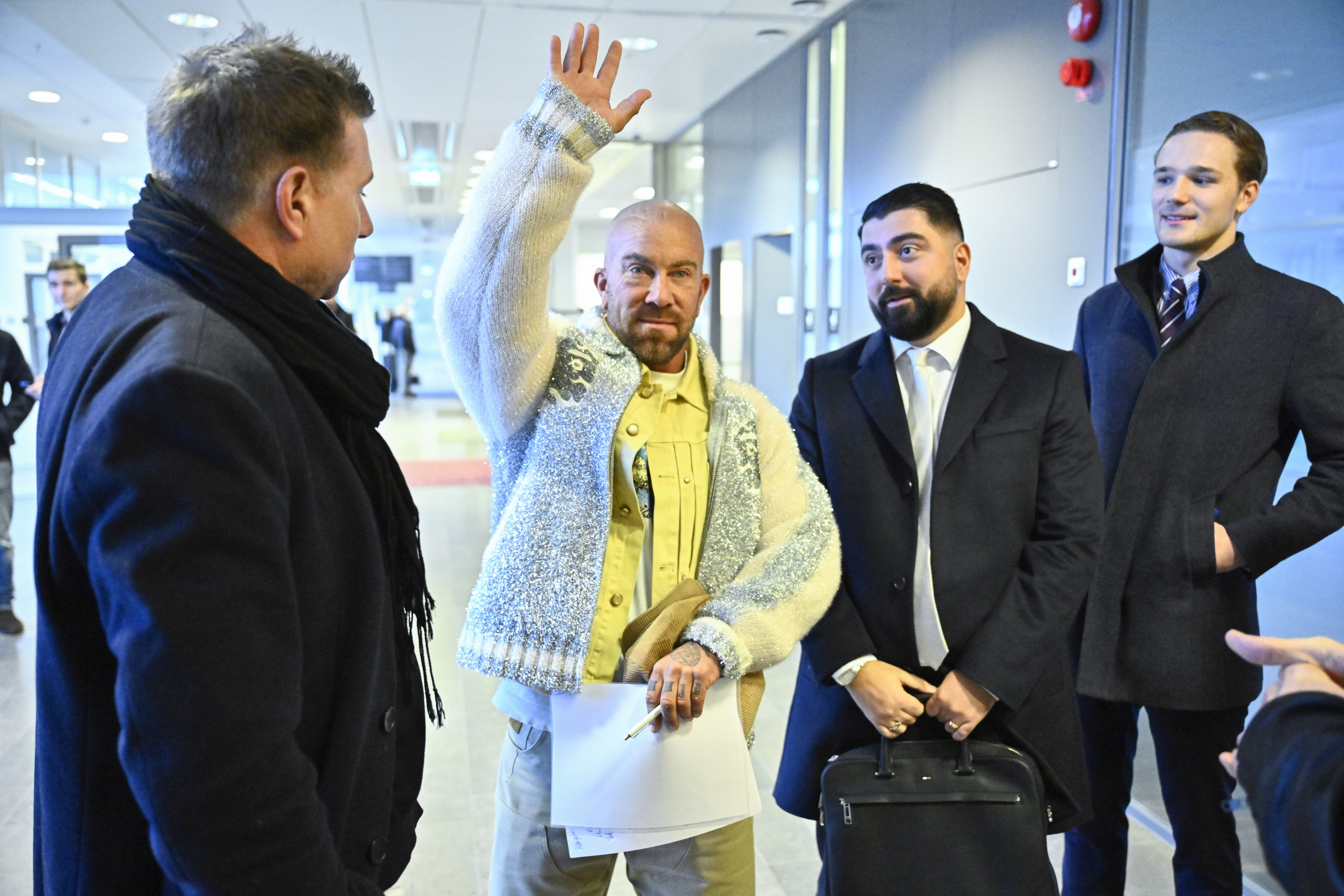Jan Emanuel i ljus tröja vinkar och håller papper. Han står med tre andra män i kostym inomhus i en modern byggnad.