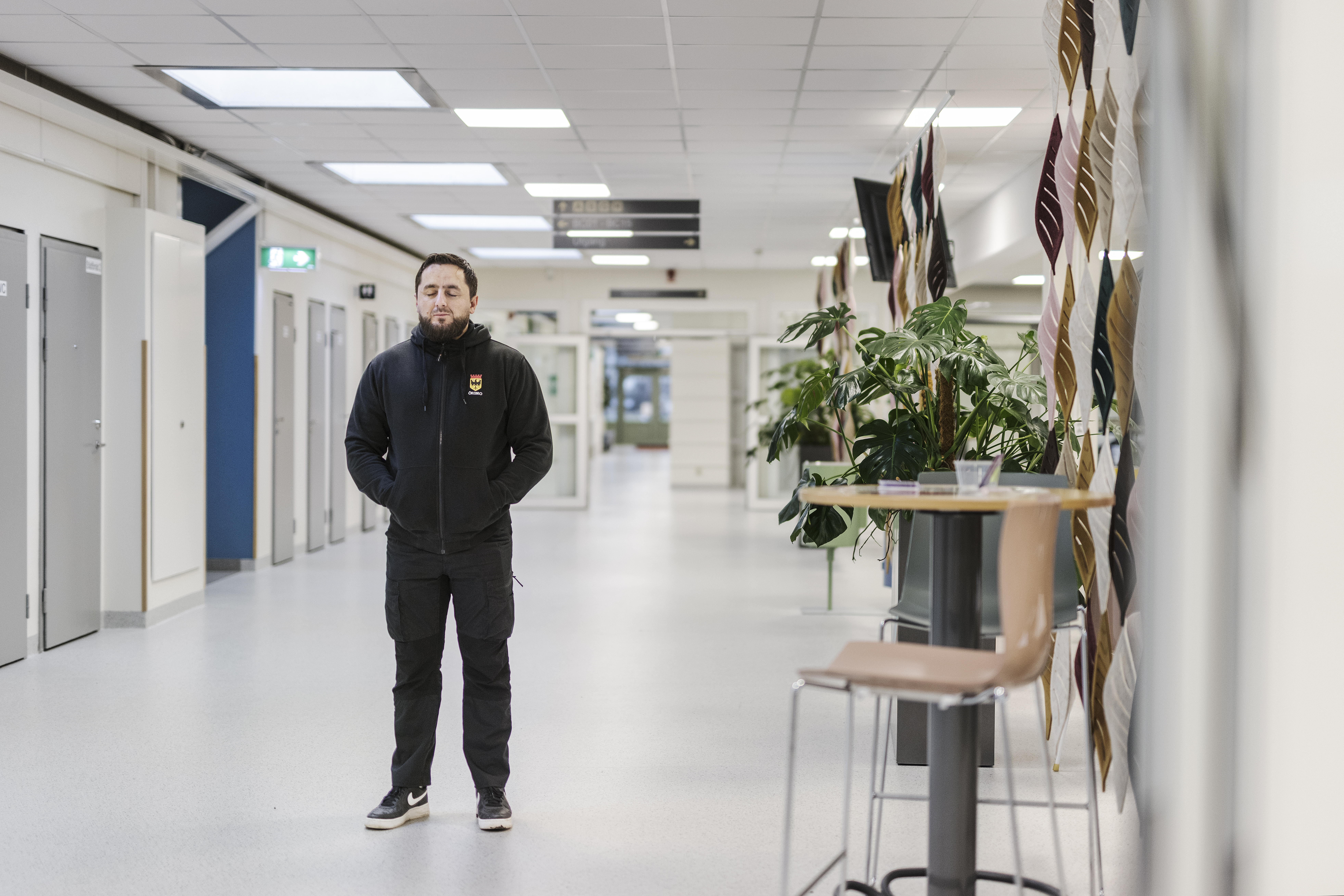 Adnan Imsirovic med svarta kläder står med ögonen slutna i en bred ljus korridor. På höger sida finns växter och ett bord med stolar.