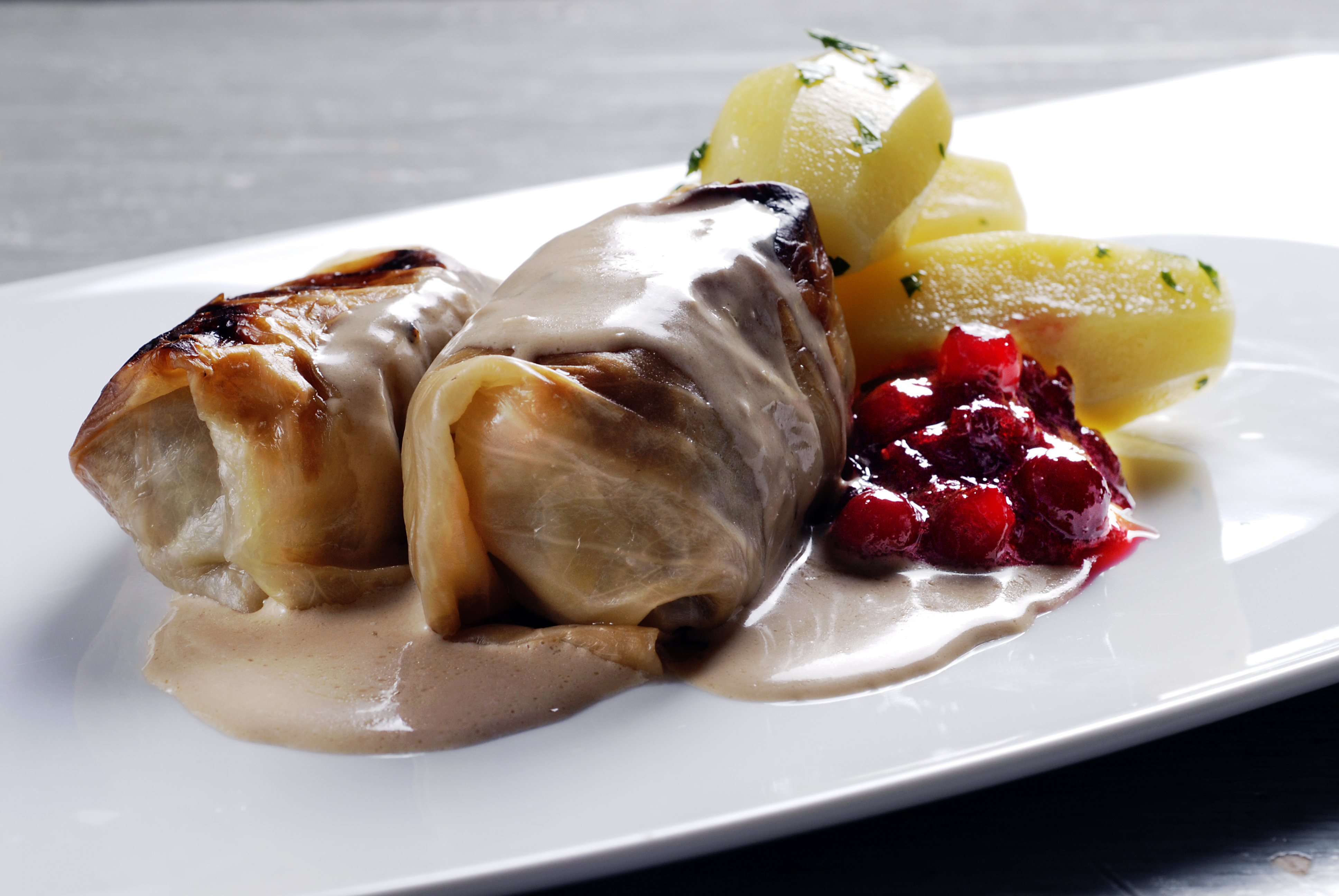 Två kåldolmar med gräddsås, kokta potatisklyftor och lingonsylt på en vit tallrik.