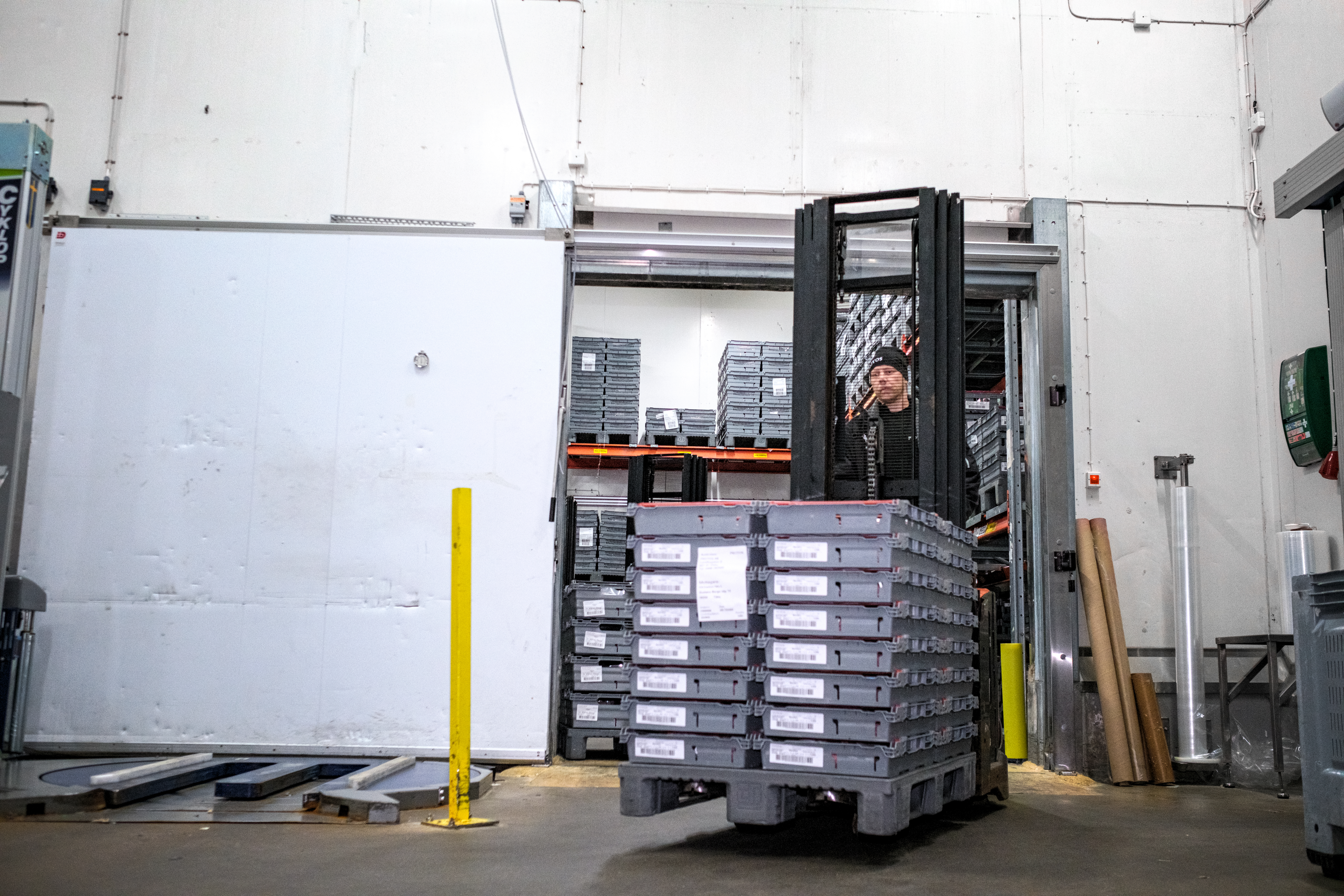 En arbetare kör en truck och flyttar en stapel metallbrickor i ett lager med vita väggar och hyllor i bakgrunden.