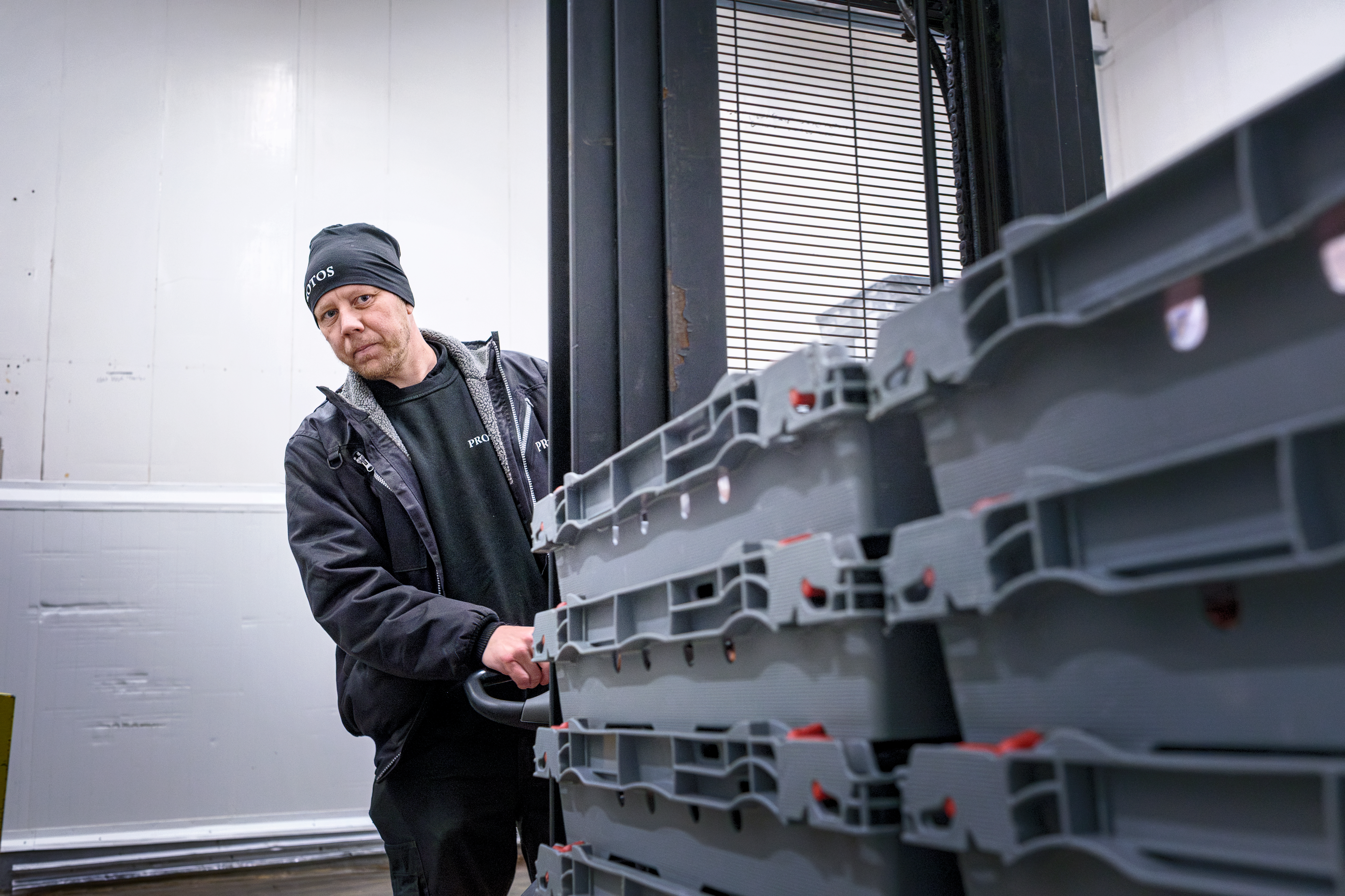 En person med svart mössa och jacka kör en lastbil och flyttar plastbehållare i en industrilokal.