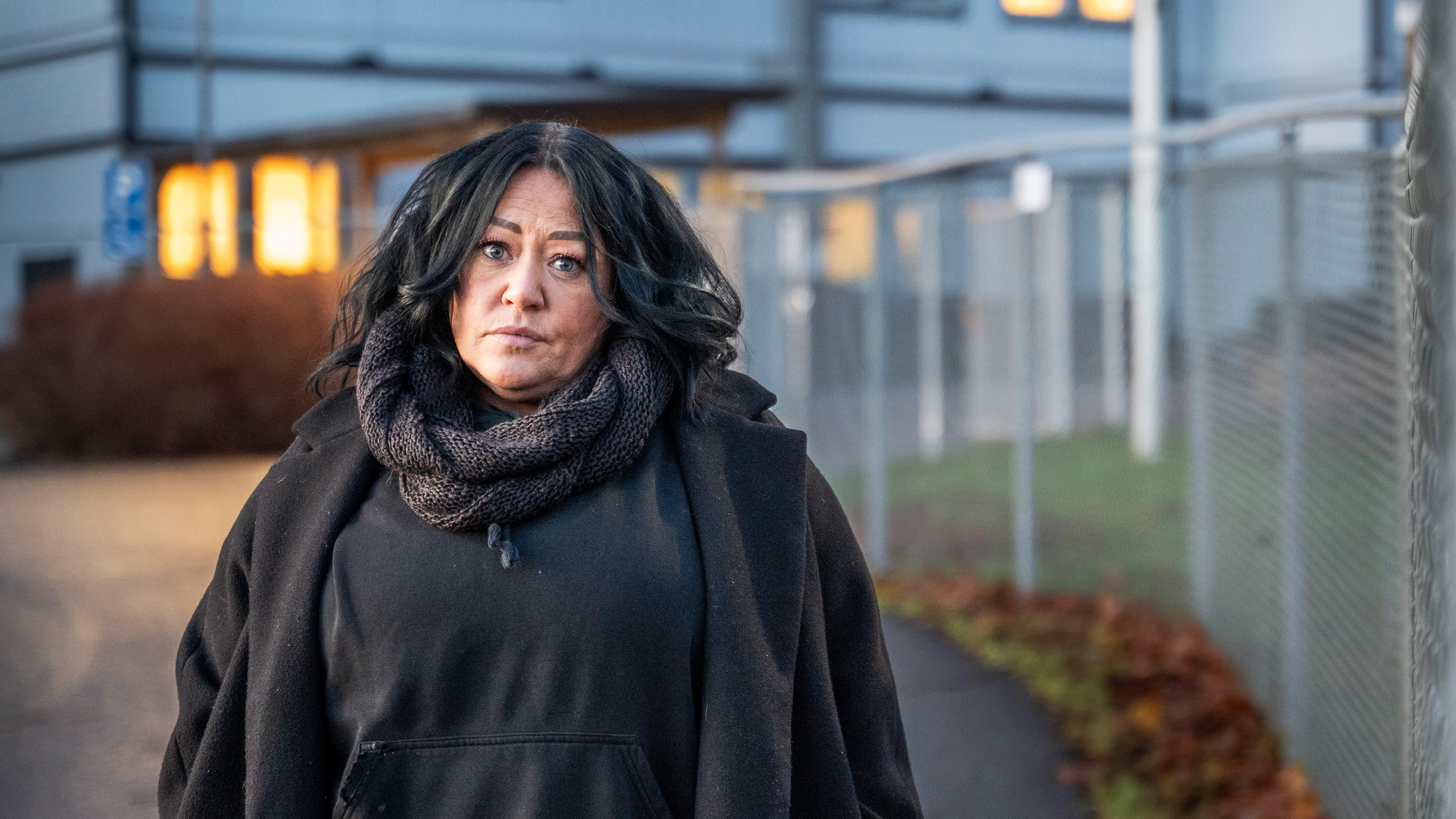 Anna Gemaius med mörkt hår och en sjal står utomhus nära ett stängsel och en byggnad, vänd lite bort från kameran.