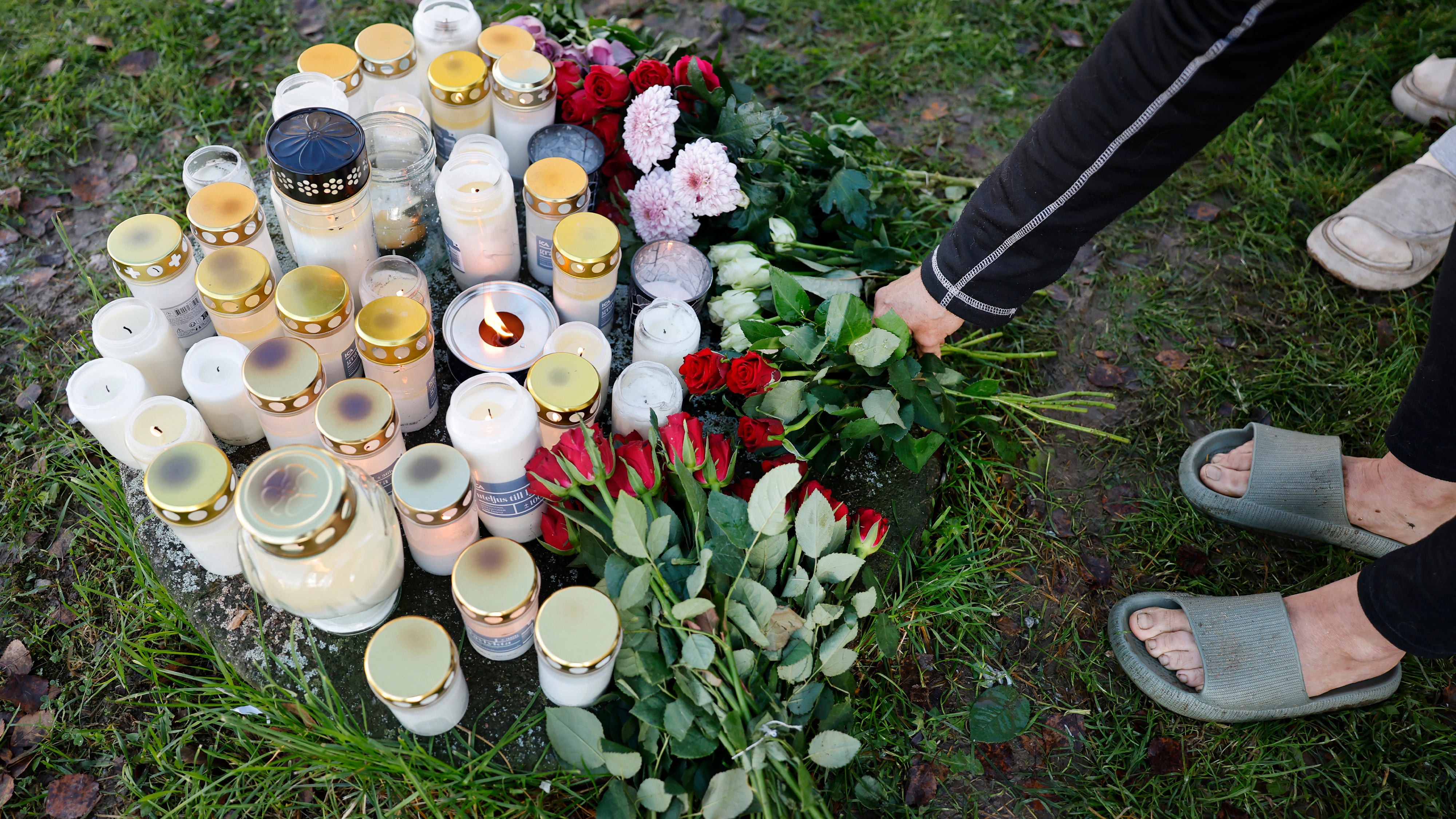 En person lägger blommor bredvid tända ljus och buketter på gräset vid en minnesplats eller hyllning.
