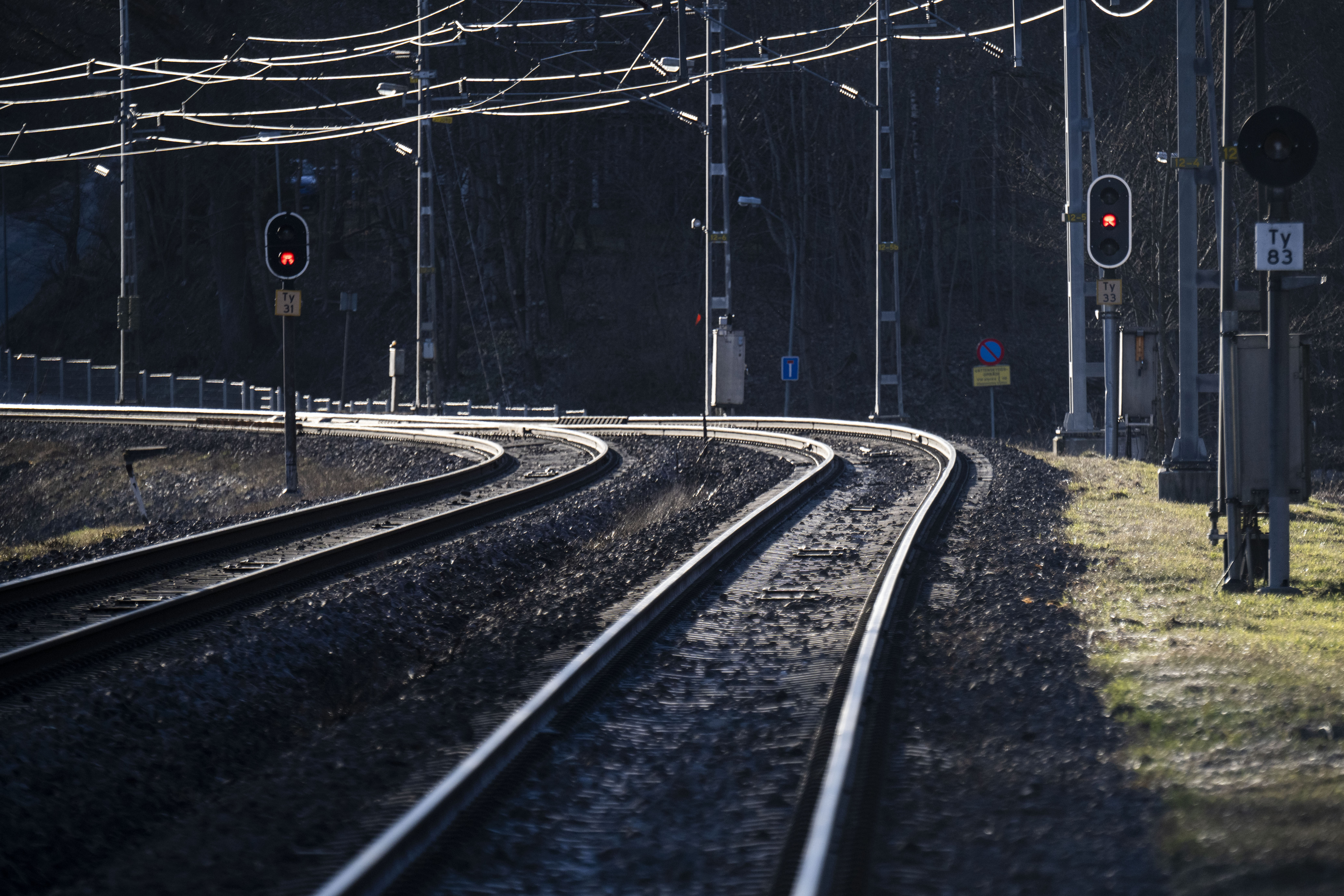 Böjda järnvägsspår med ledningar ovanför och röda signaler, fotograferade under dagen med träd i bakgrunden.