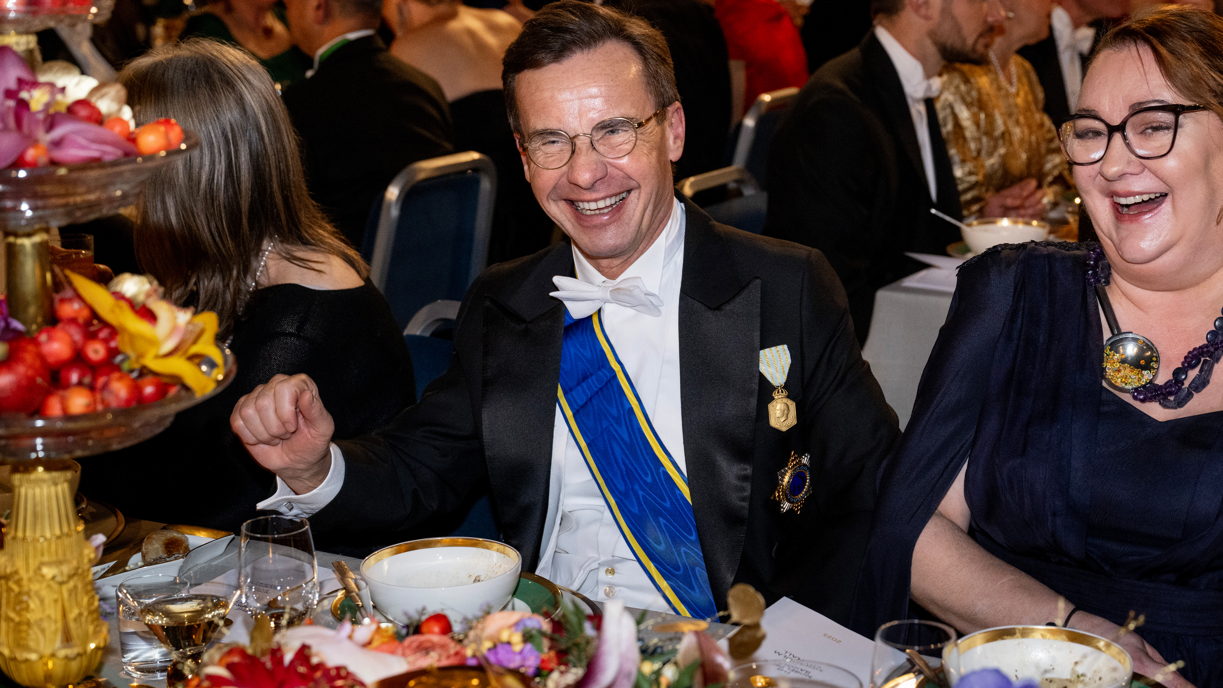 En man i högtidsdräkt med blå ordensband och medaljer sitter vid ett bankettbord tillsammans med andra gäster och mat.