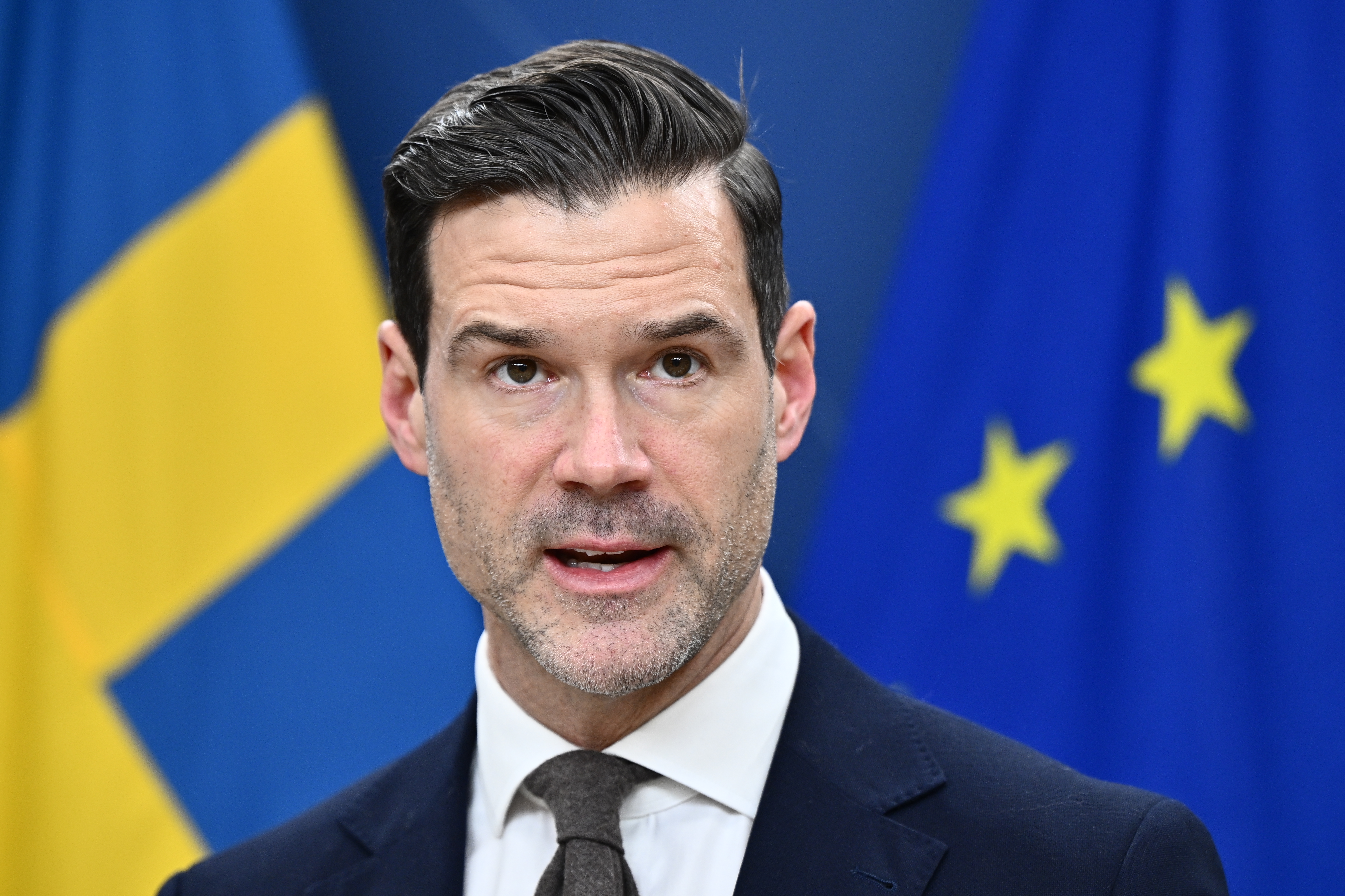 En man i kostym talar framför Sveriges och Europeiska unionens flaggor.