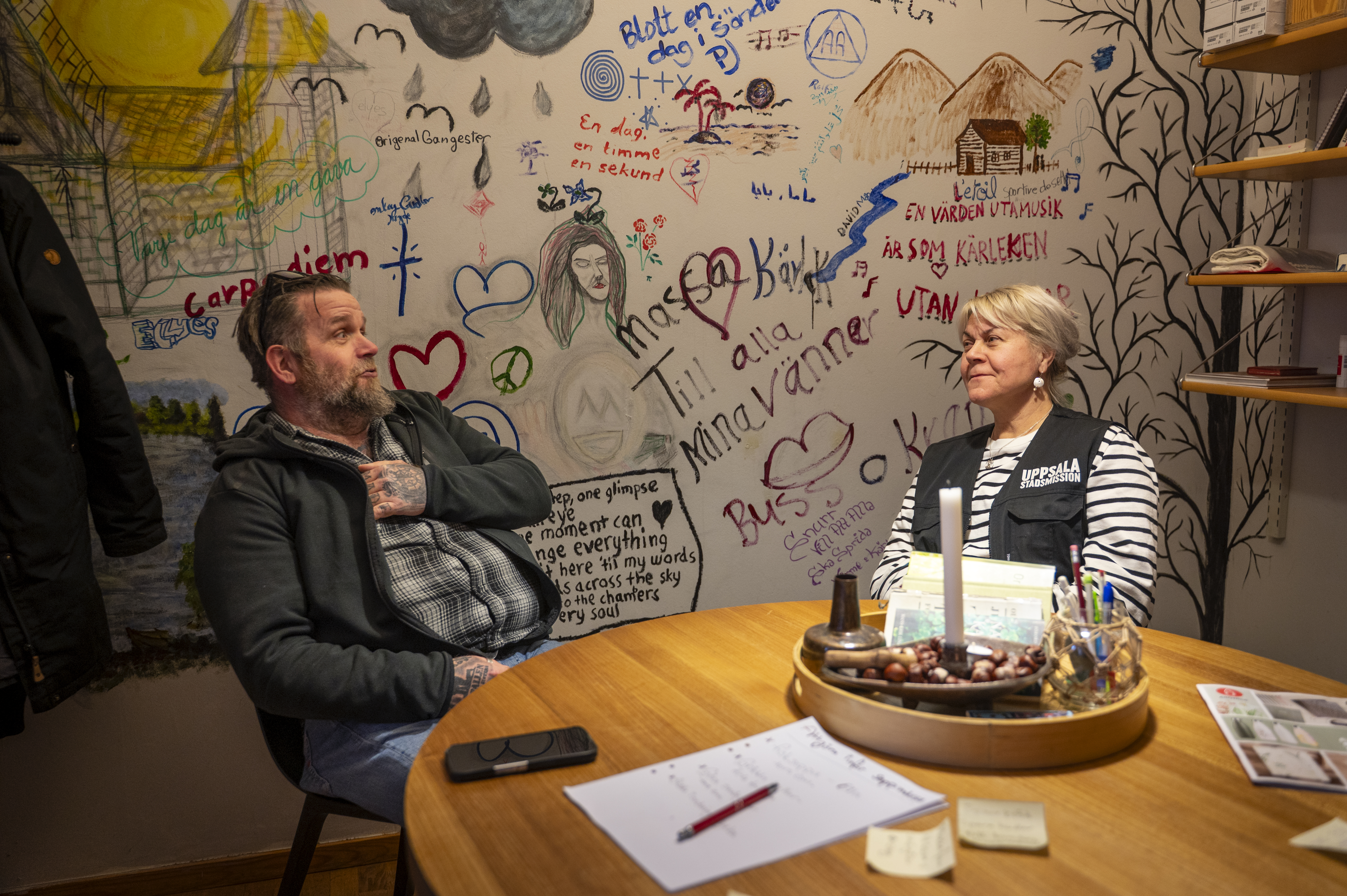 En man och en kvinna sitter vid ett bord i ett rum med teckningar och texter på väggen; papper och kontorsmaterial ligger på bordet.