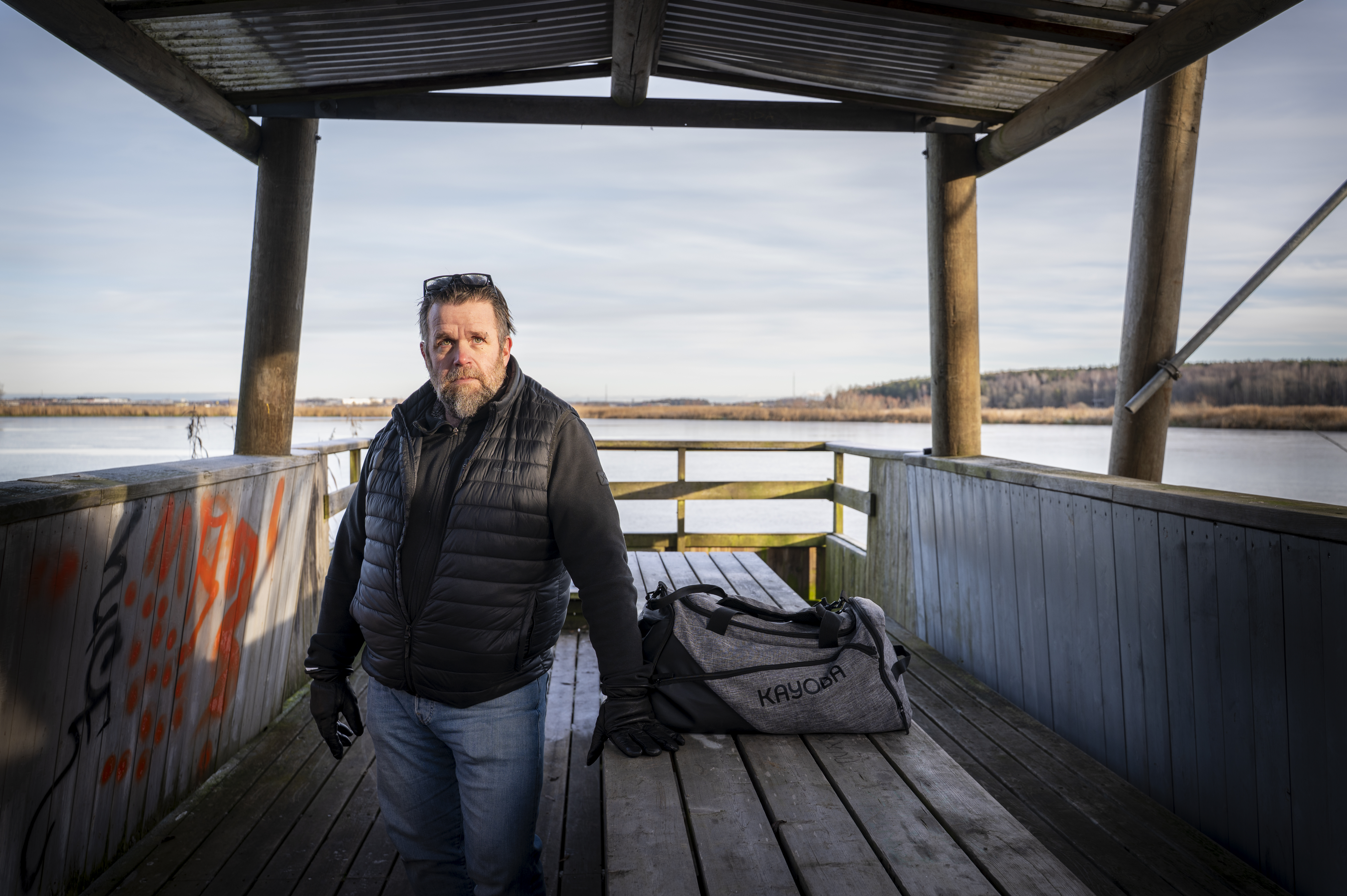 En man med svart väst och handskar står på en brygga vid vattnet bredvid en grå sportväska med texten "KRYOOR".