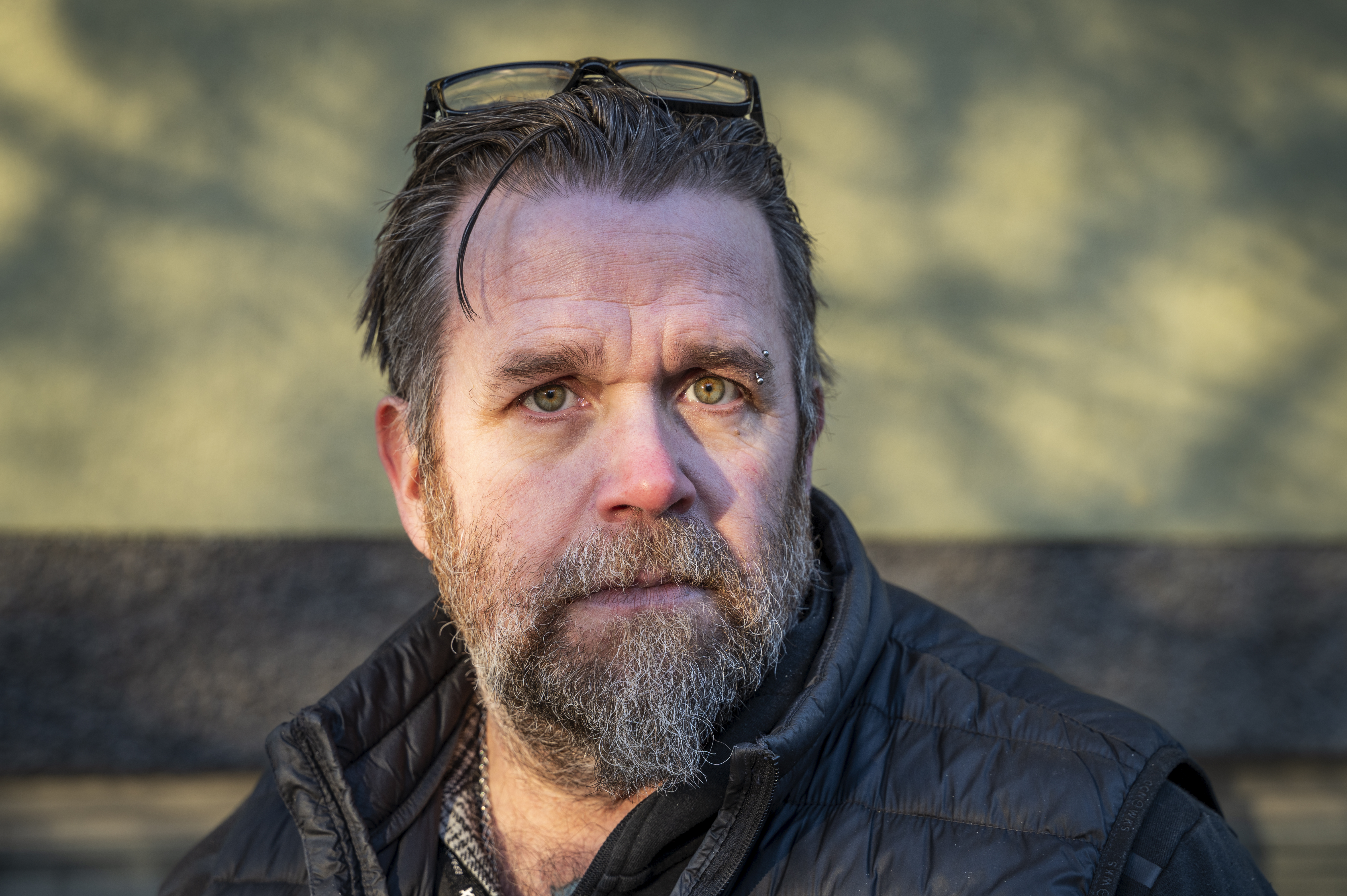 En man med grått skägg och mustasch, glasögon på huvudet och svart väst står utomhus med oskarp bakgrund.
