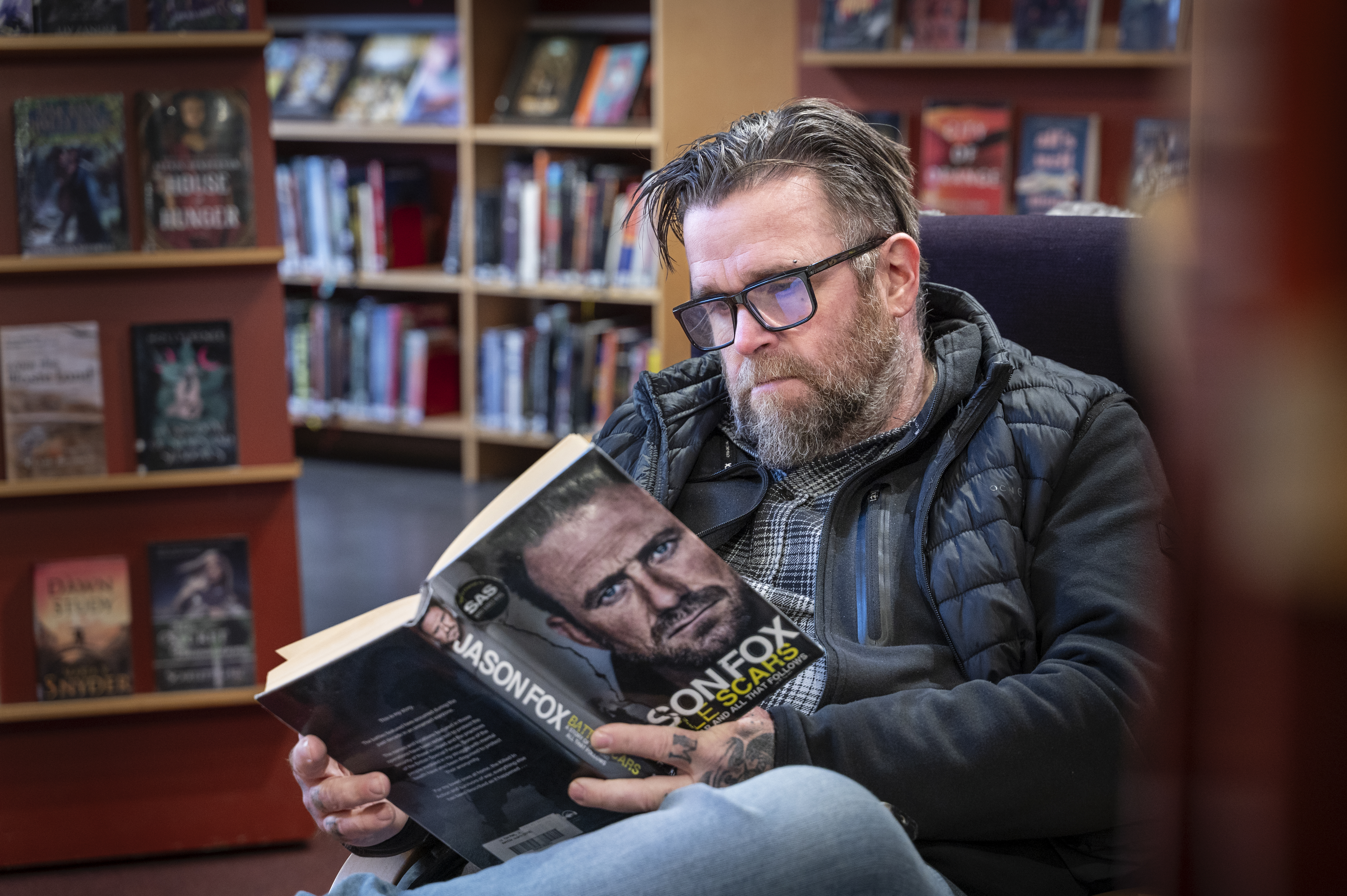 En man med glasögon och skägg sitter i ett bibliotek och läser boken "Jason Fox: Livet under eld". Bokhyllor syns bakom.