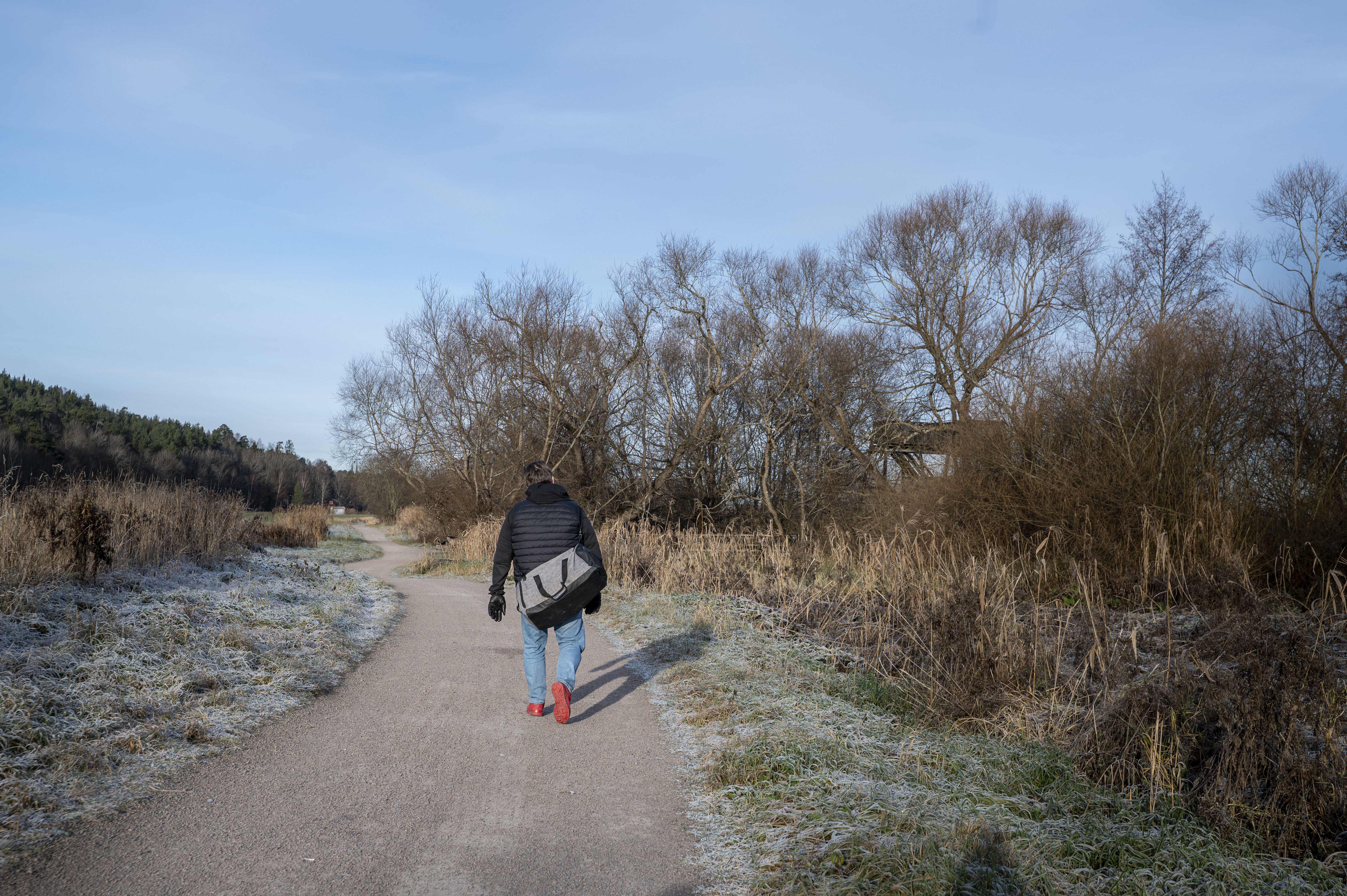 En person med svart jacka och röda skor går ensam på en isig landsväg med torrt gräs och träd utan löv under blå himmel.