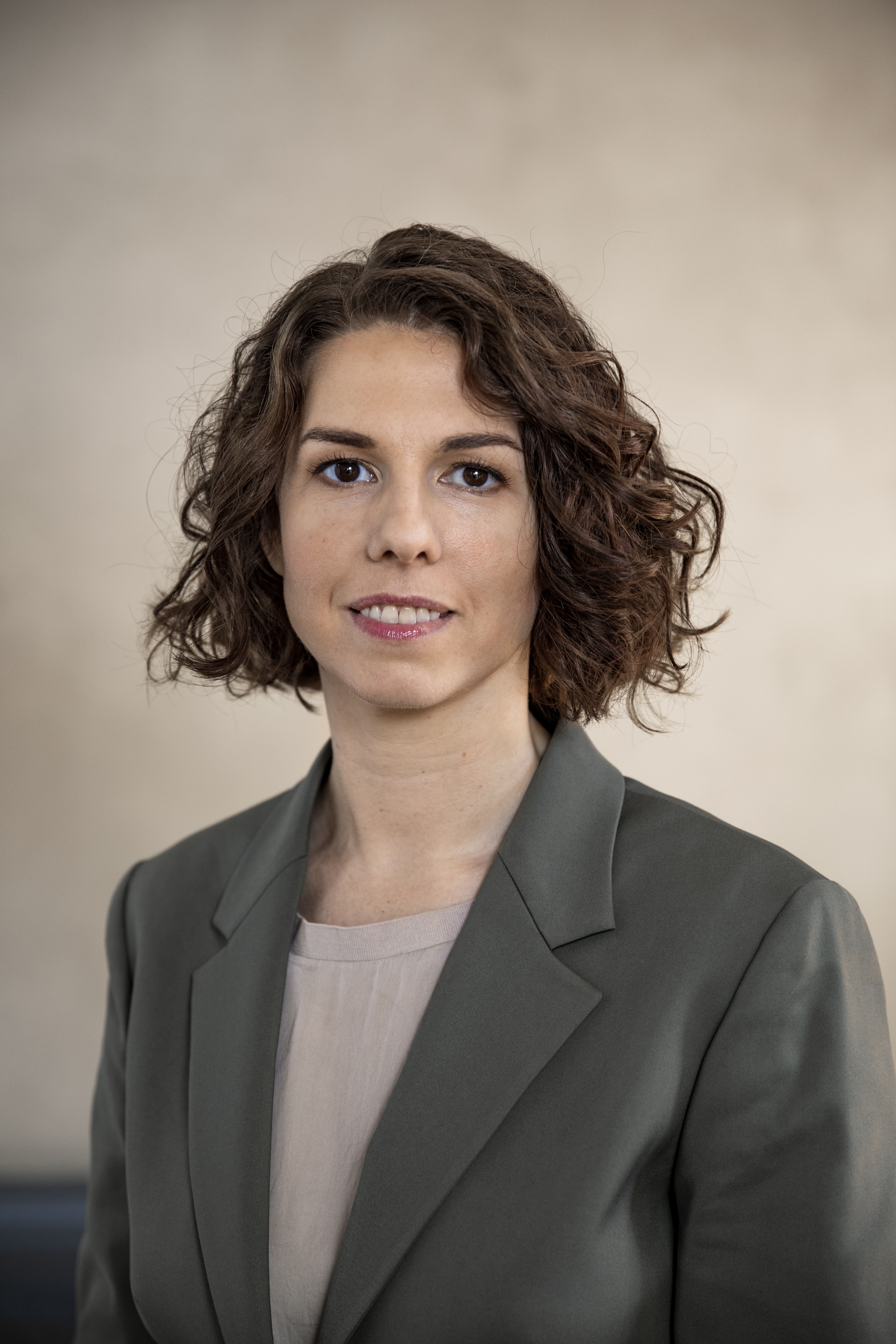 En kvinna med kort, lockigt brunt hår har på sig en grå kavaj och en beige topp och står mot en ljus bakgrund.