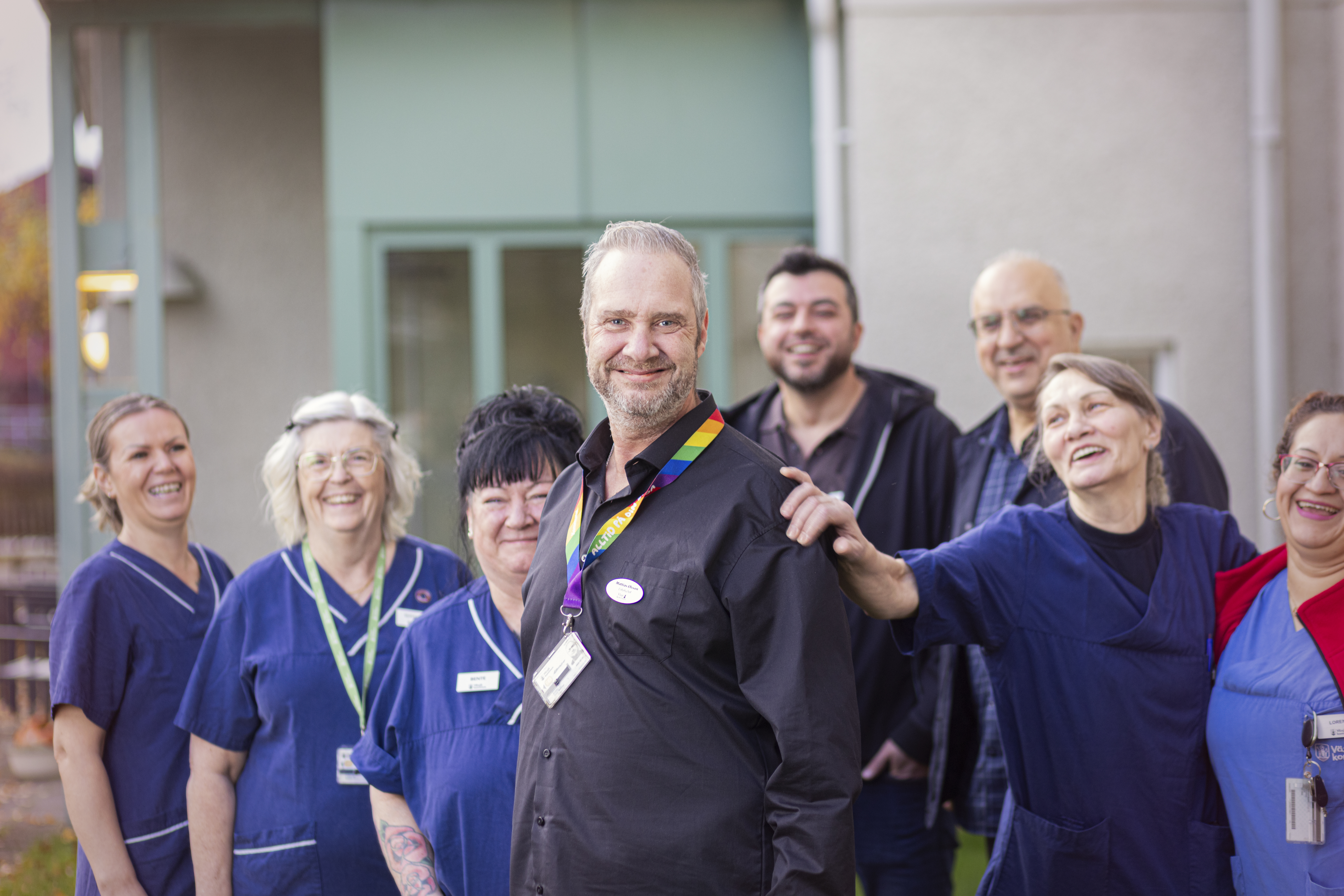 En grupp sjukvårdspersonal är samlad utanför byggnaden, några bär arbetsuniformer. Matthias Olsson längst fram bär en regnbågsnyckelring.