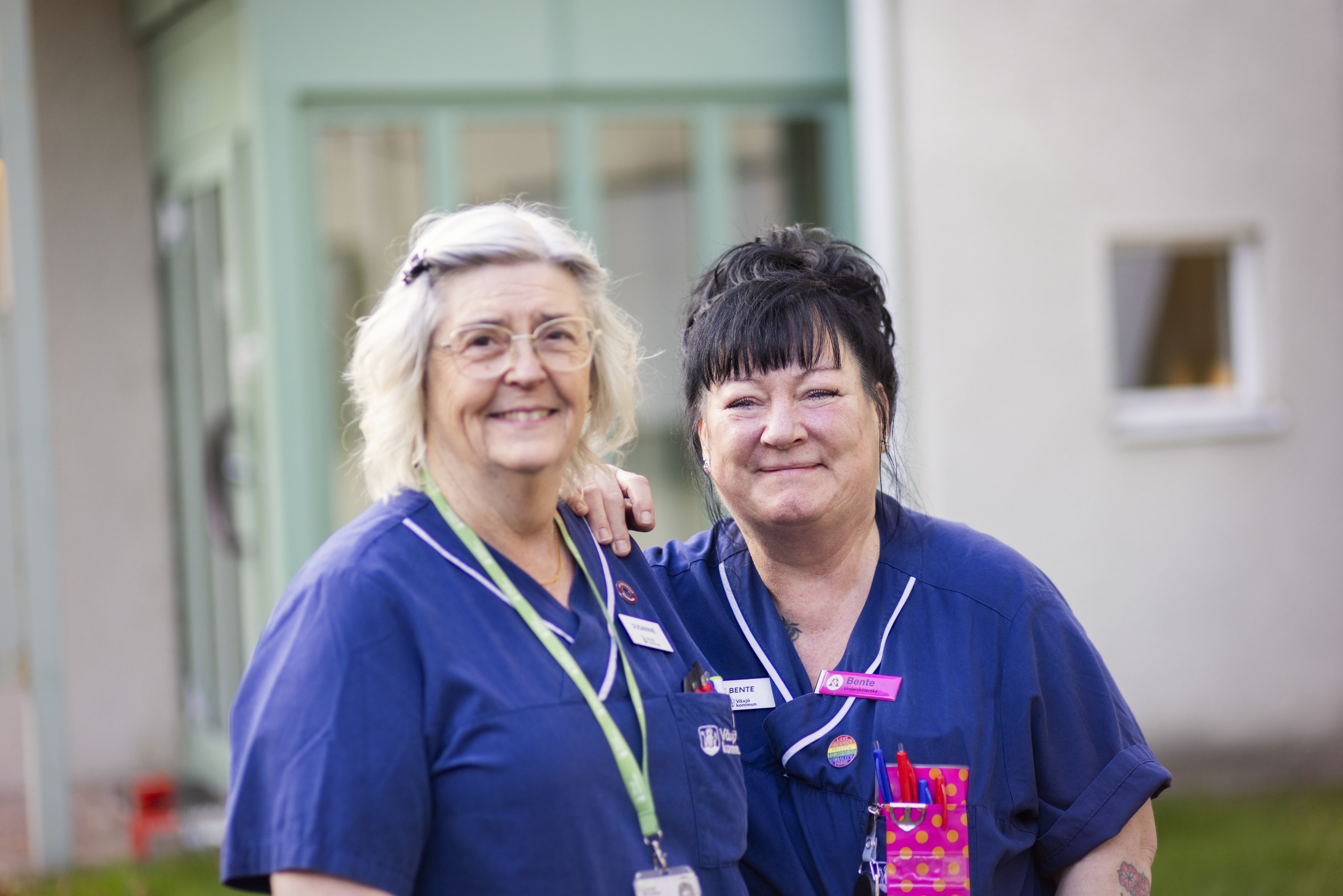 Två kvinnor i blåa kläder står utanför en byggnad och tittar in i kameran.