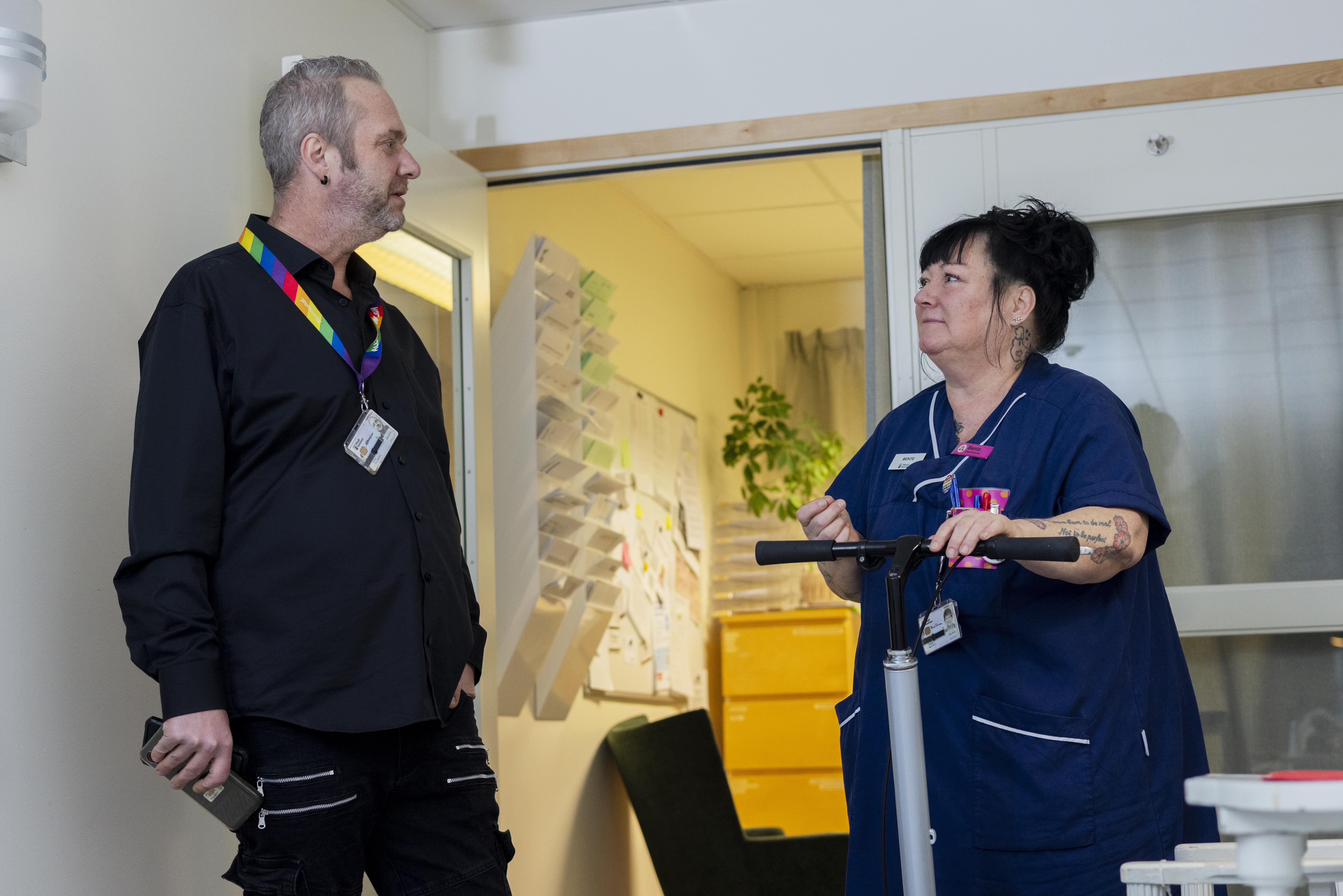 En man i svarta kläder pratar med en kvinna i blå vårdkläder som håller i ett gånghjälpmedel i en vårdmiljö.