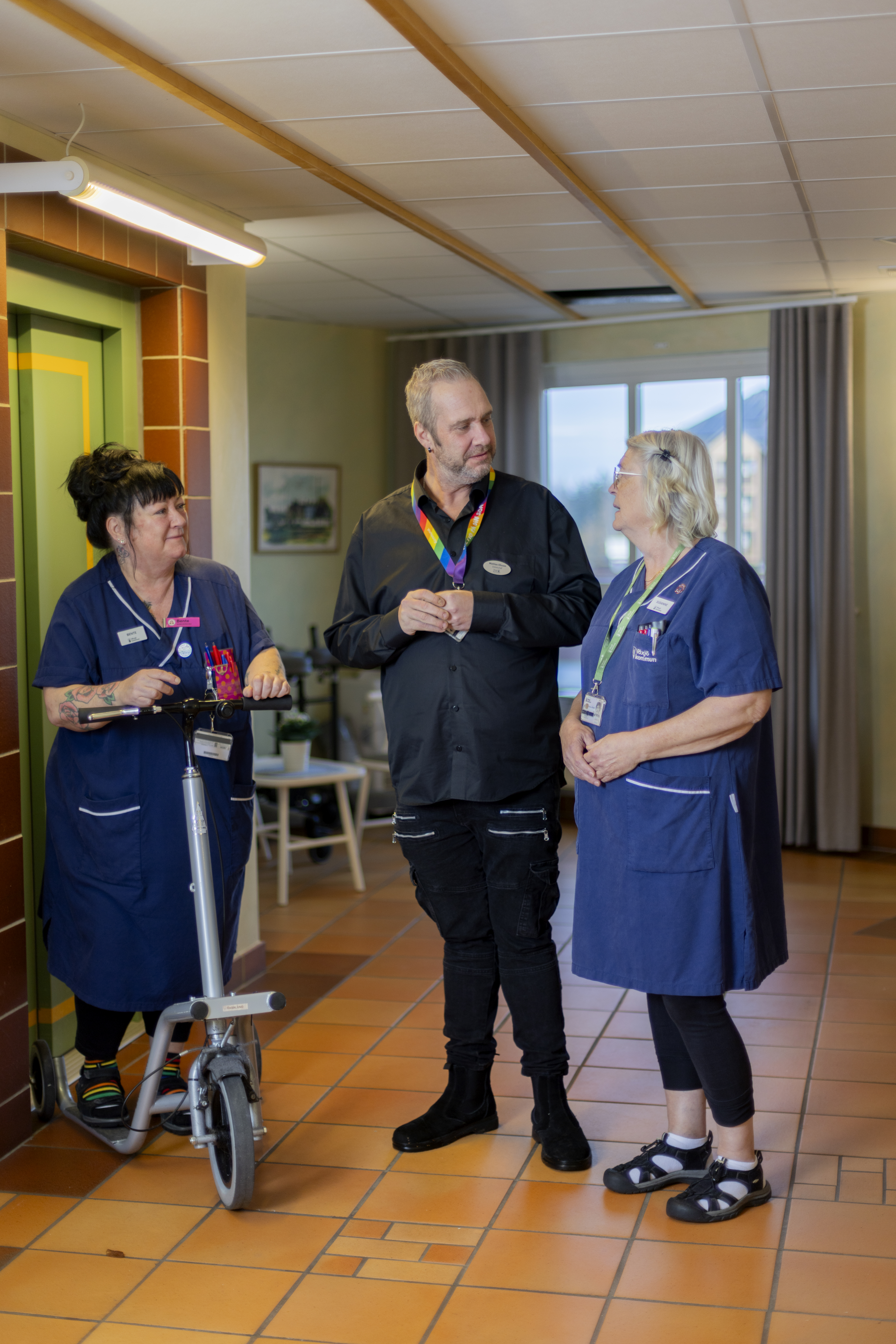 Tre vuxna, två i blå uniformer och en i svart, står och pratar i en hall med klinkergolv. En person håller i en liten elscooter.