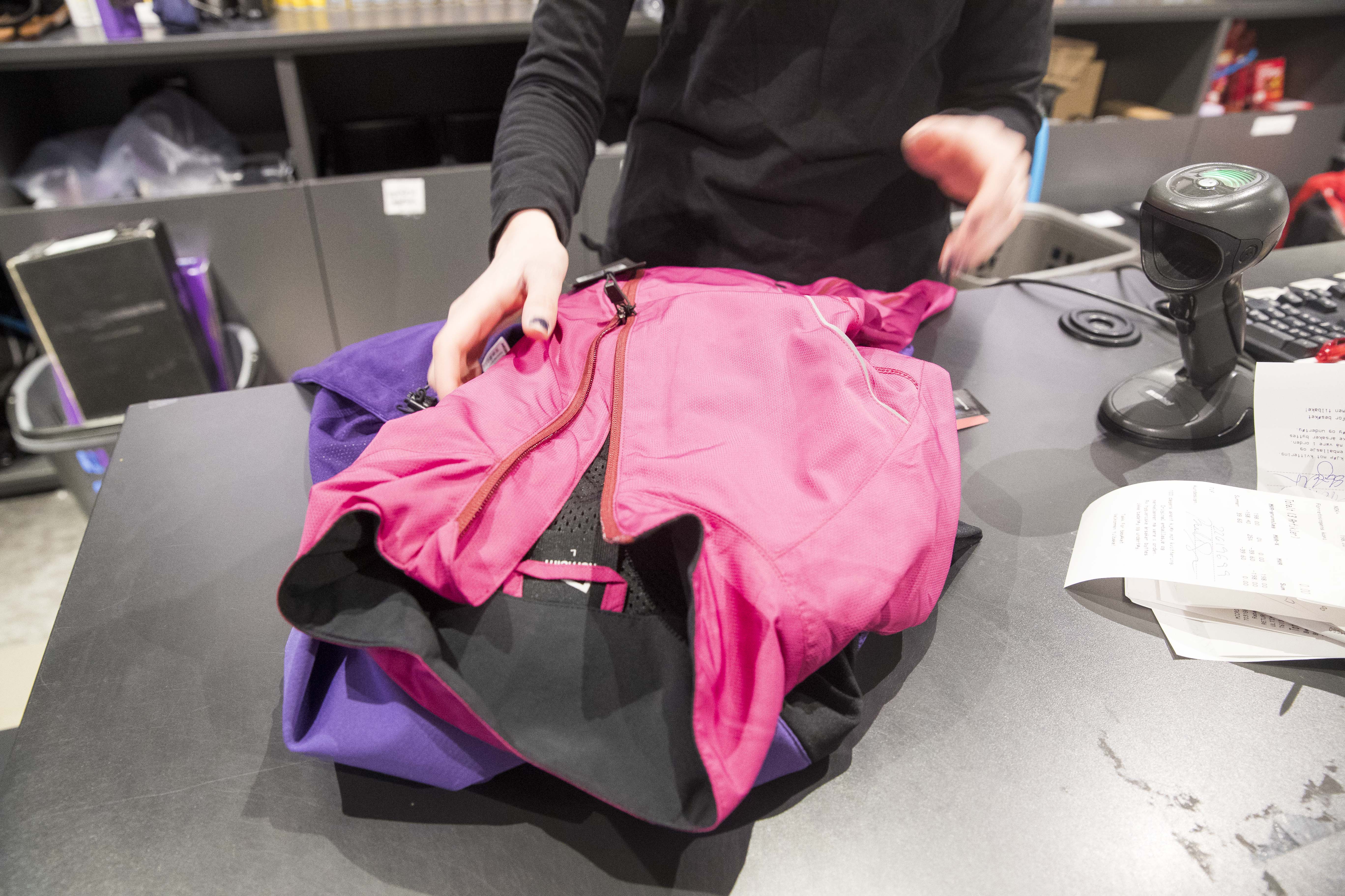 En person i svarta kläder hanterar en retur eller ett byte av en rosa och lila jacka vid en butikskassa med kvitto och streckkodsläsare.