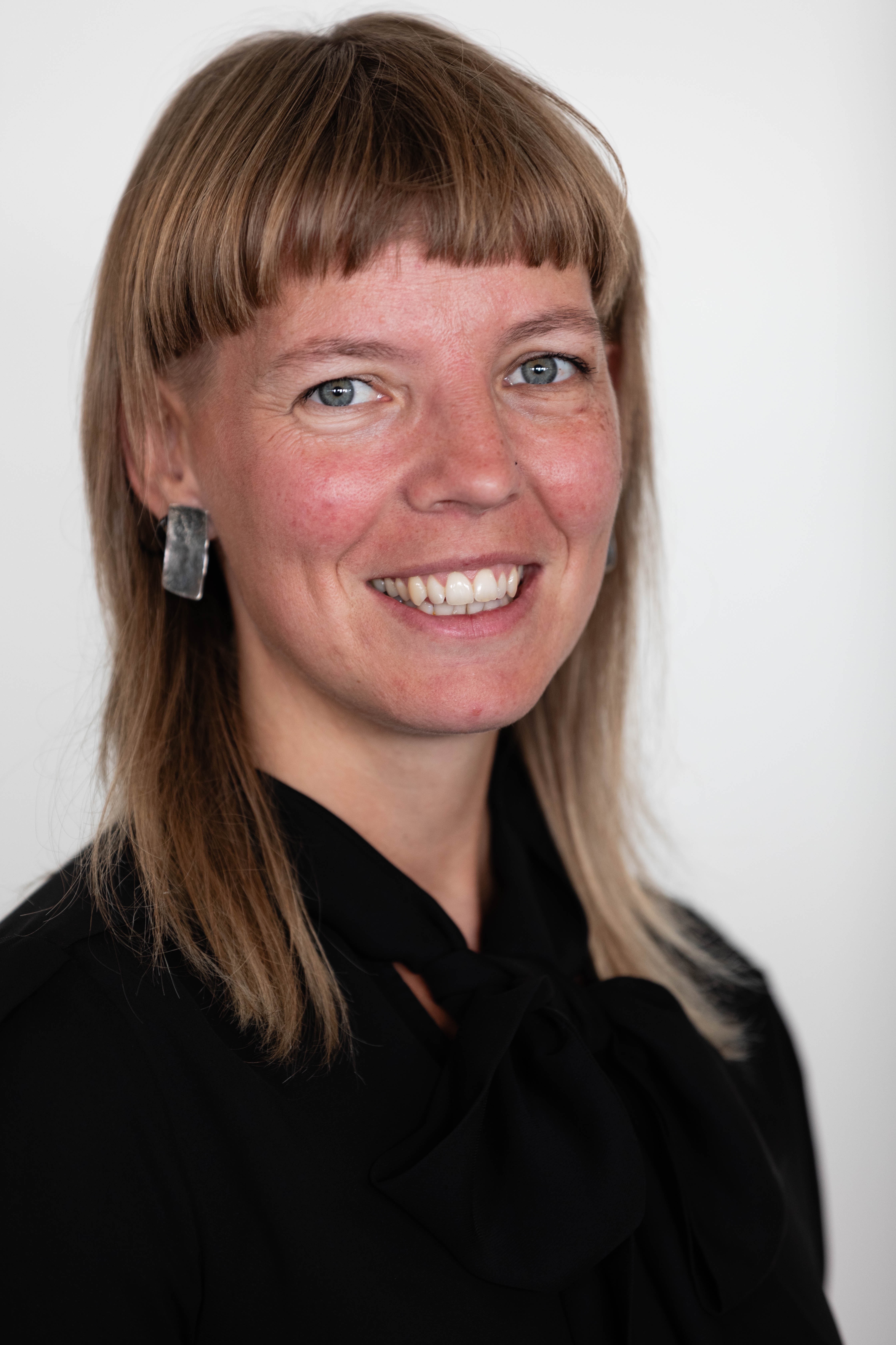 En kvinna med rakt, ljusbrunt hår och lugg har på sig en svart topp med rosett och geometriska örhängen mot vit bakgrund.