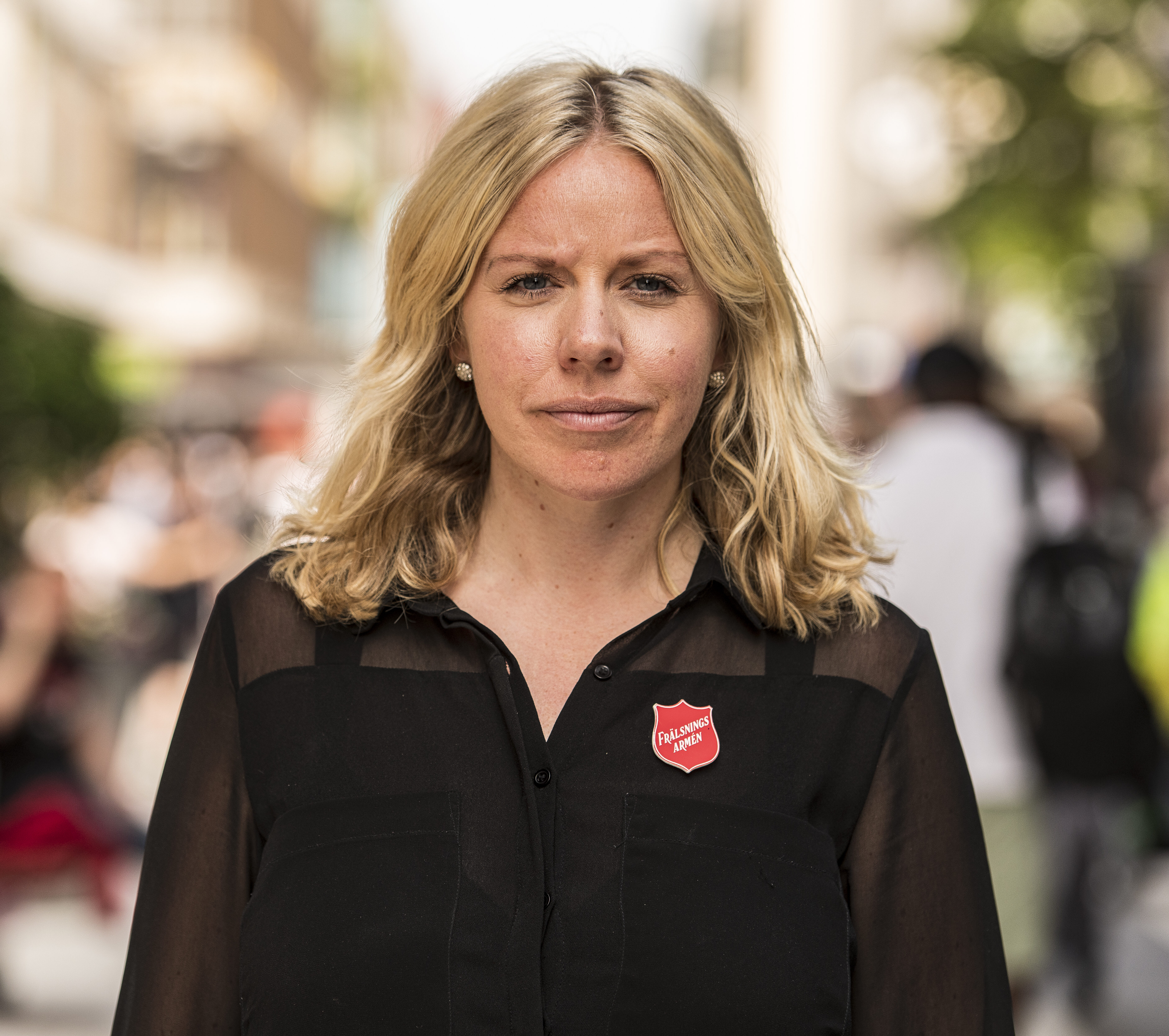 En kvinna med blont hår, svart tröja och en Frälsningsarmén-nål står utomhus med en suddig gata i bakgrunden.