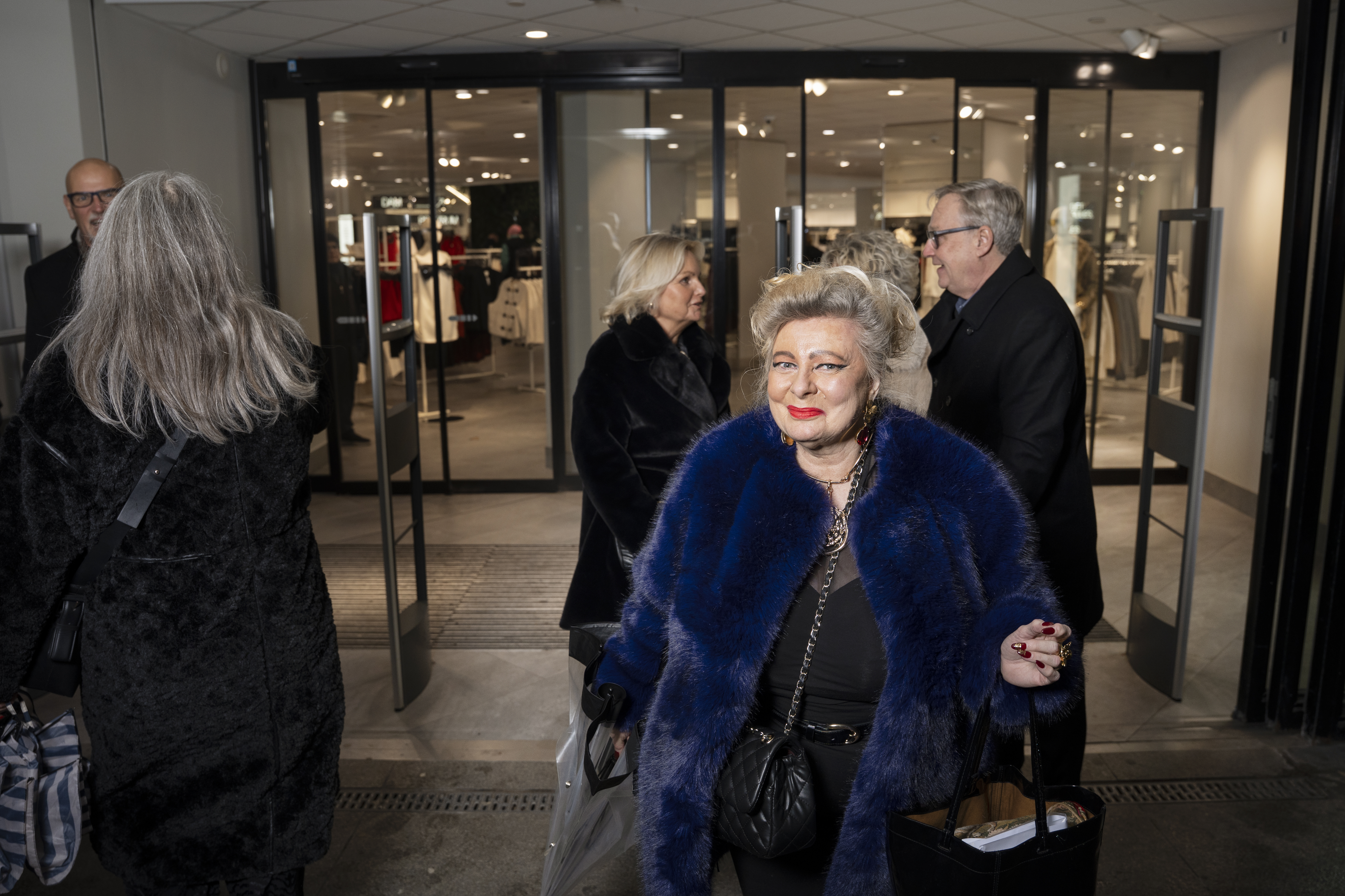 En kvinna i blå pälsjacka bär shoppingpåsar och lämnar en klädbutik tillsammans med andra personer.
