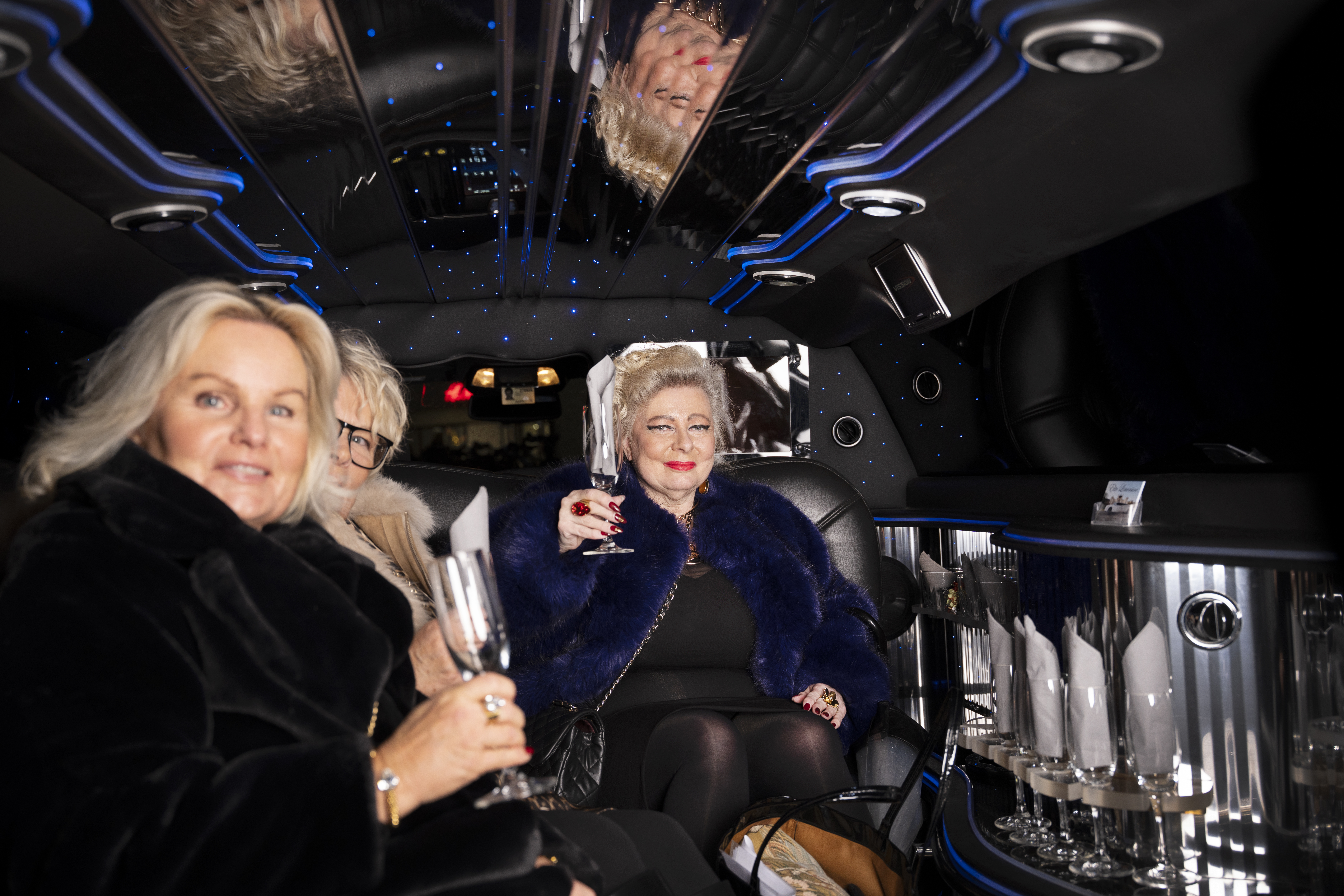 Tre kvinnor sitter i baksätet av en limousine med tomma glas, spegeltak och blått ljus syns. En kvinna håller upp ett glas och ler mot kameran.