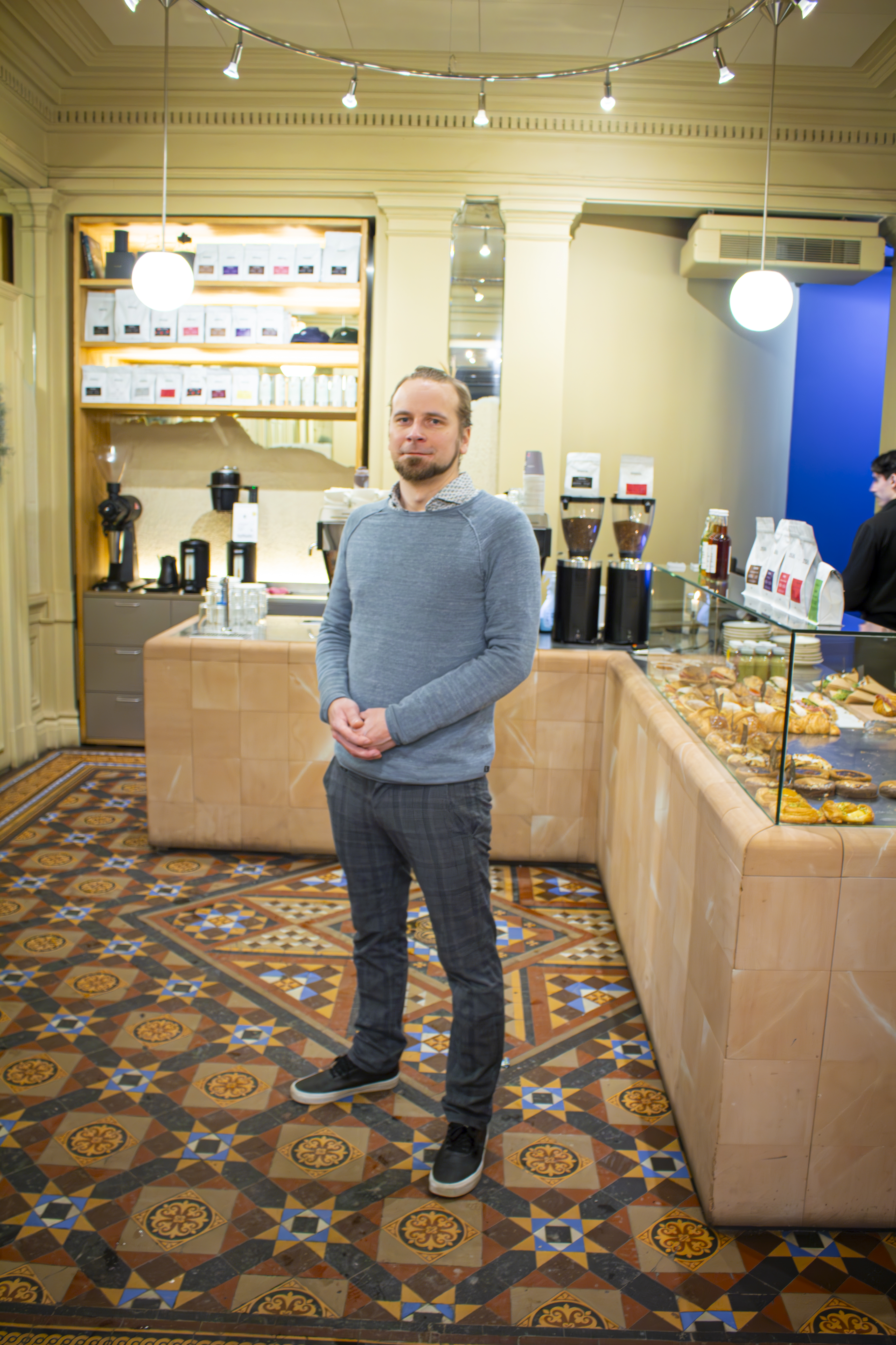 En man står i ett bageri eller café med bakverk i en glasmonter och kaffemaskiner på bänken bakom honom.