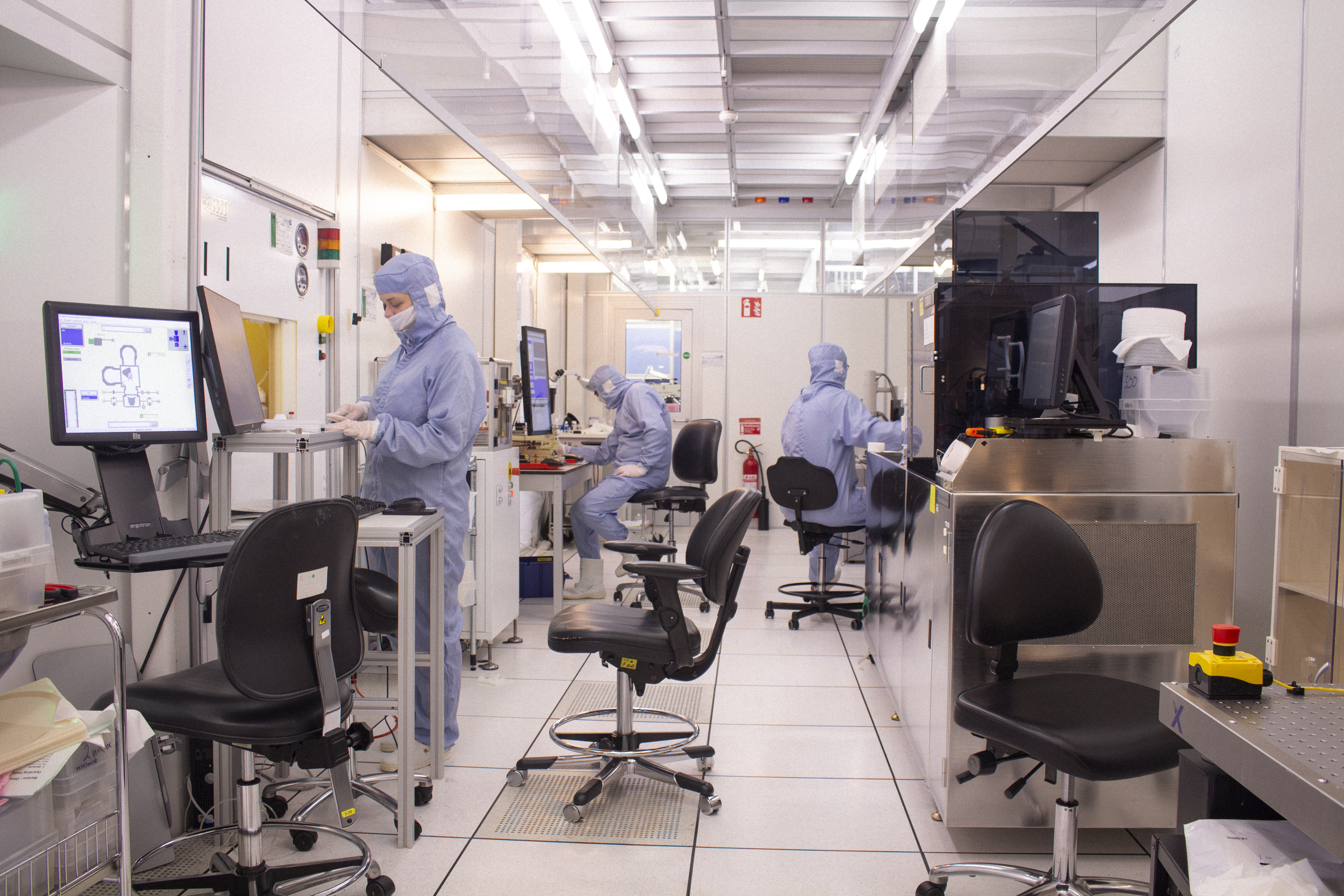 Forskare i skyddsdräkter arbetar vid datorer och labbstationer i ett renrumslabb med vita väggar och utrustning.