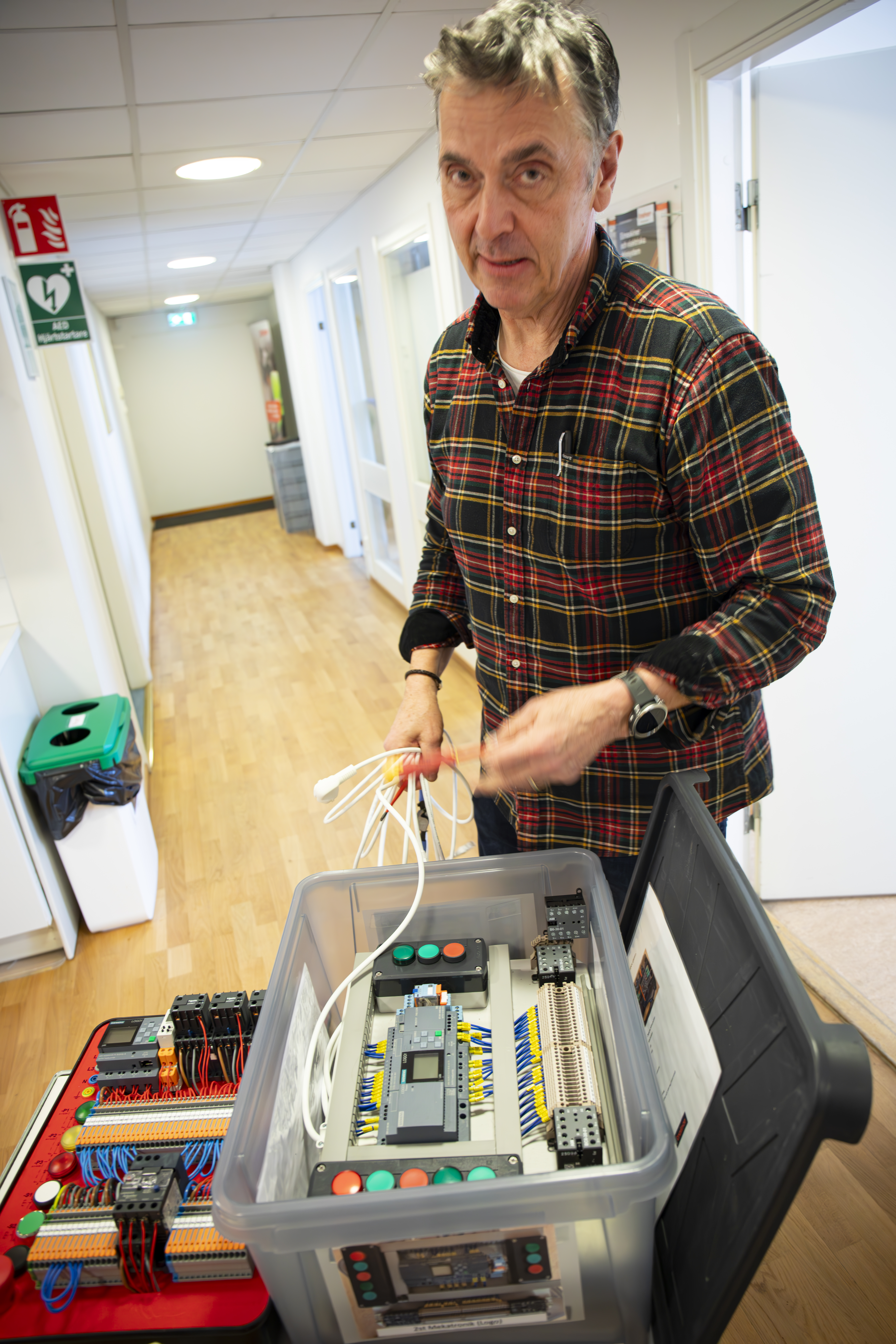 En man står i en hall bredvid en plastlåda med elektroniska komponenter och sladdar, och håller flera kablar i handen.