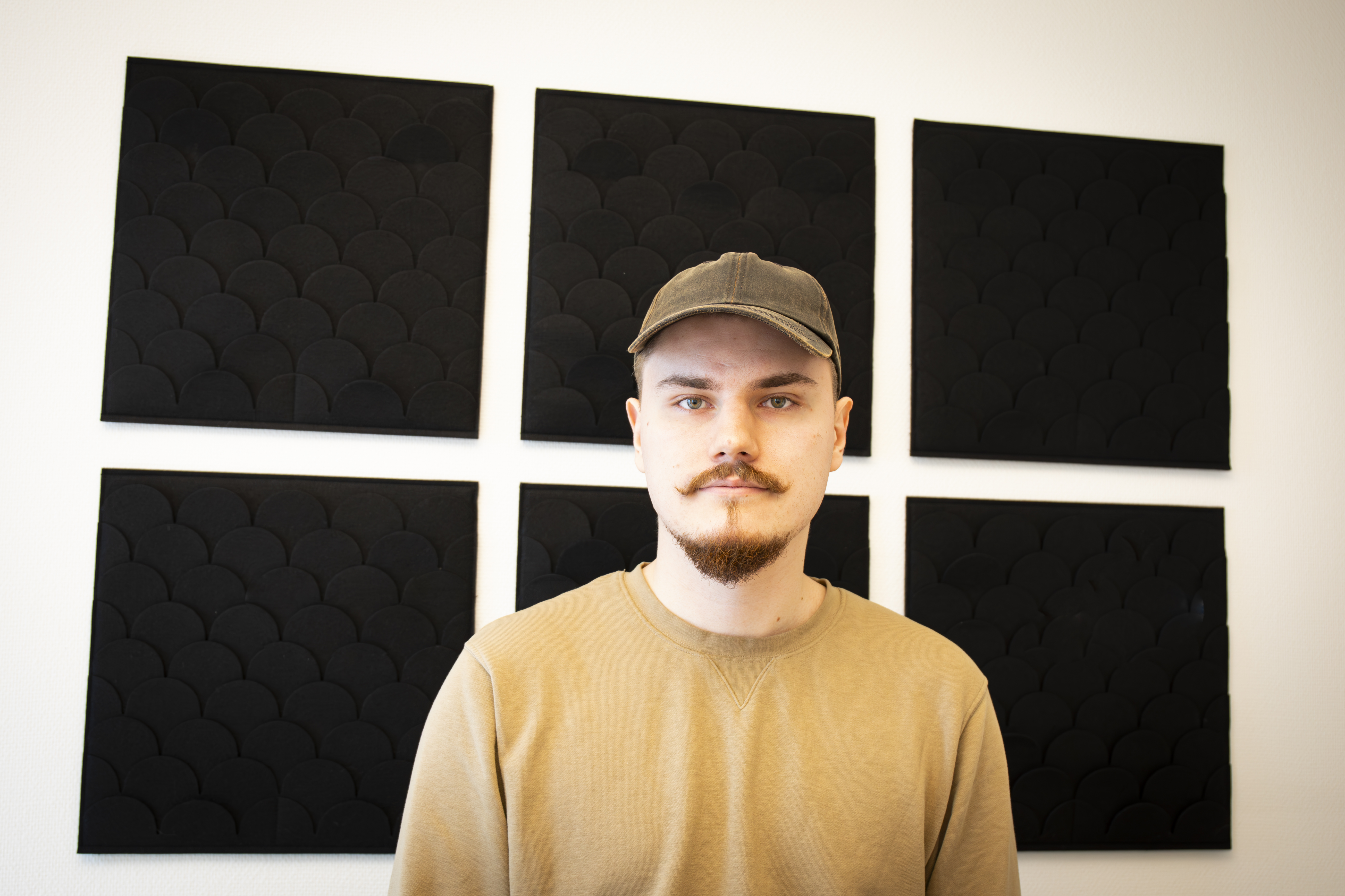 En man med beige tröja och keps står framför en vit vägg med fem svarta fyrkantiga paneler.