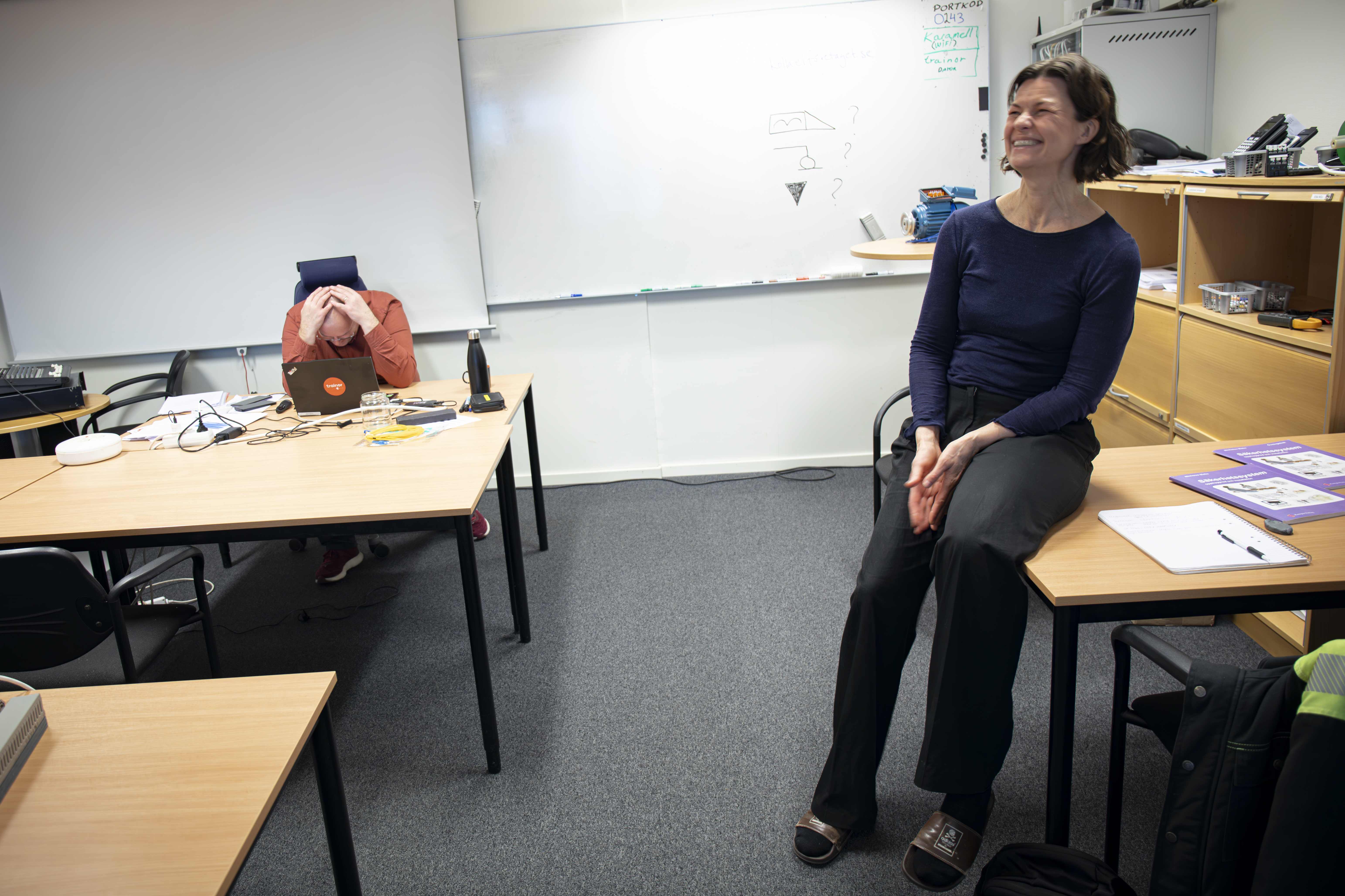 En kvinna sitter på ett skrivbord och ler, medan en annan person sitter vid ett bord med huvudet i händerna i ett klassrum med whiteboard.