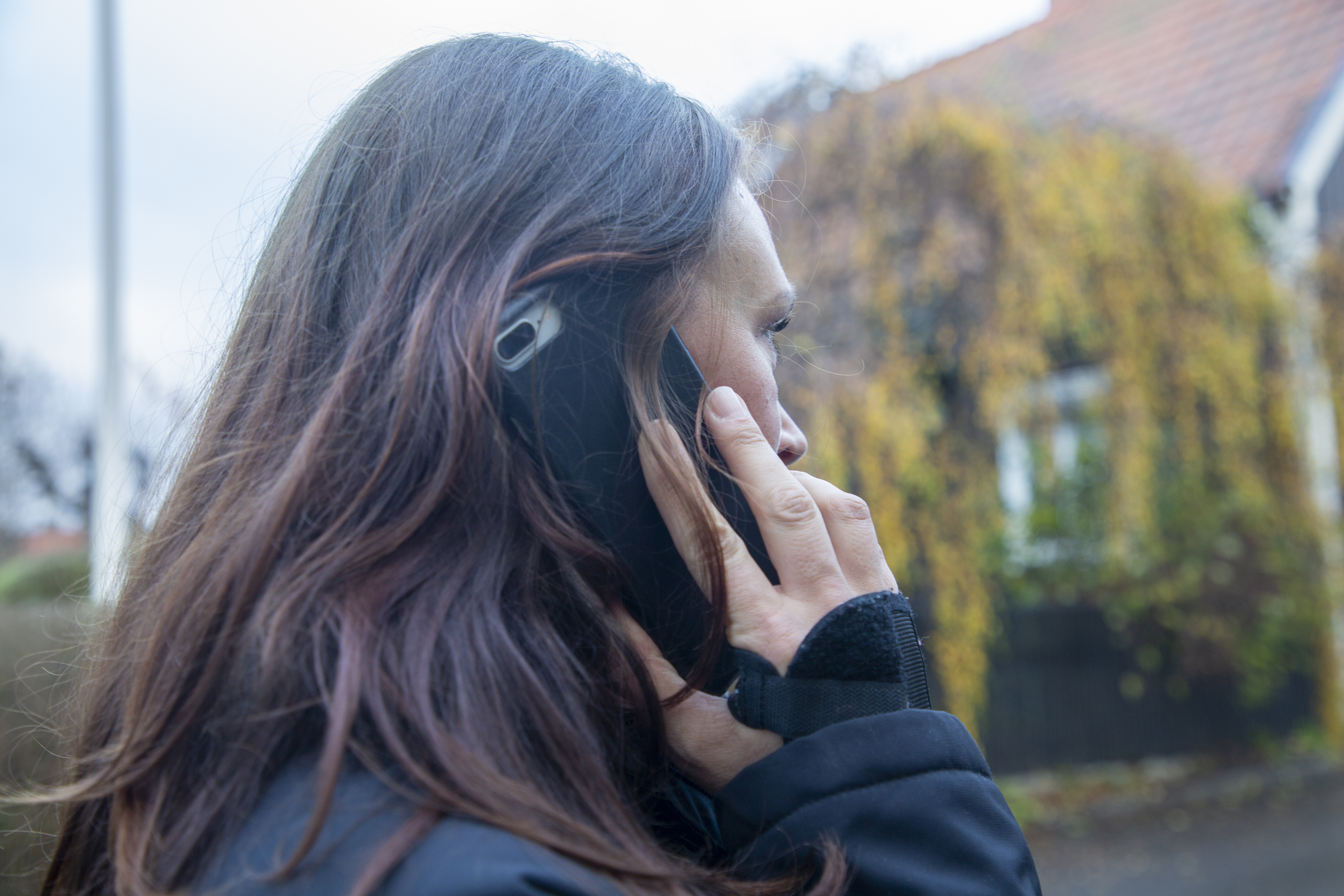 En person med långt brunt hår håller en smartphone mot örat och pratar i telefon utomhus, med ett hus och växter i bakgrunden.