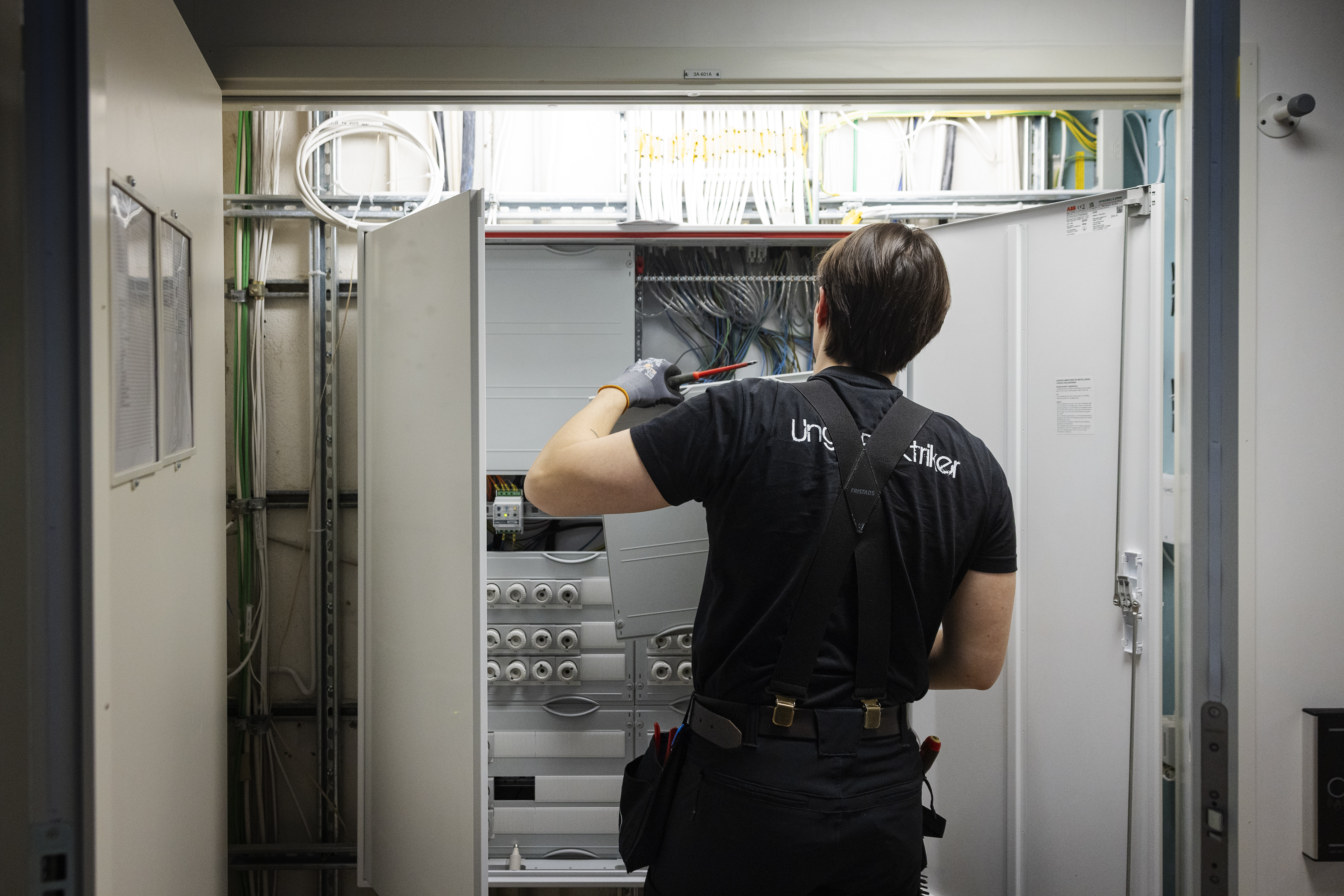 En person med svarta arbetskläder och handskar använder ett verktyg för att undersöka eller reparera elutrustning i ett skåp.