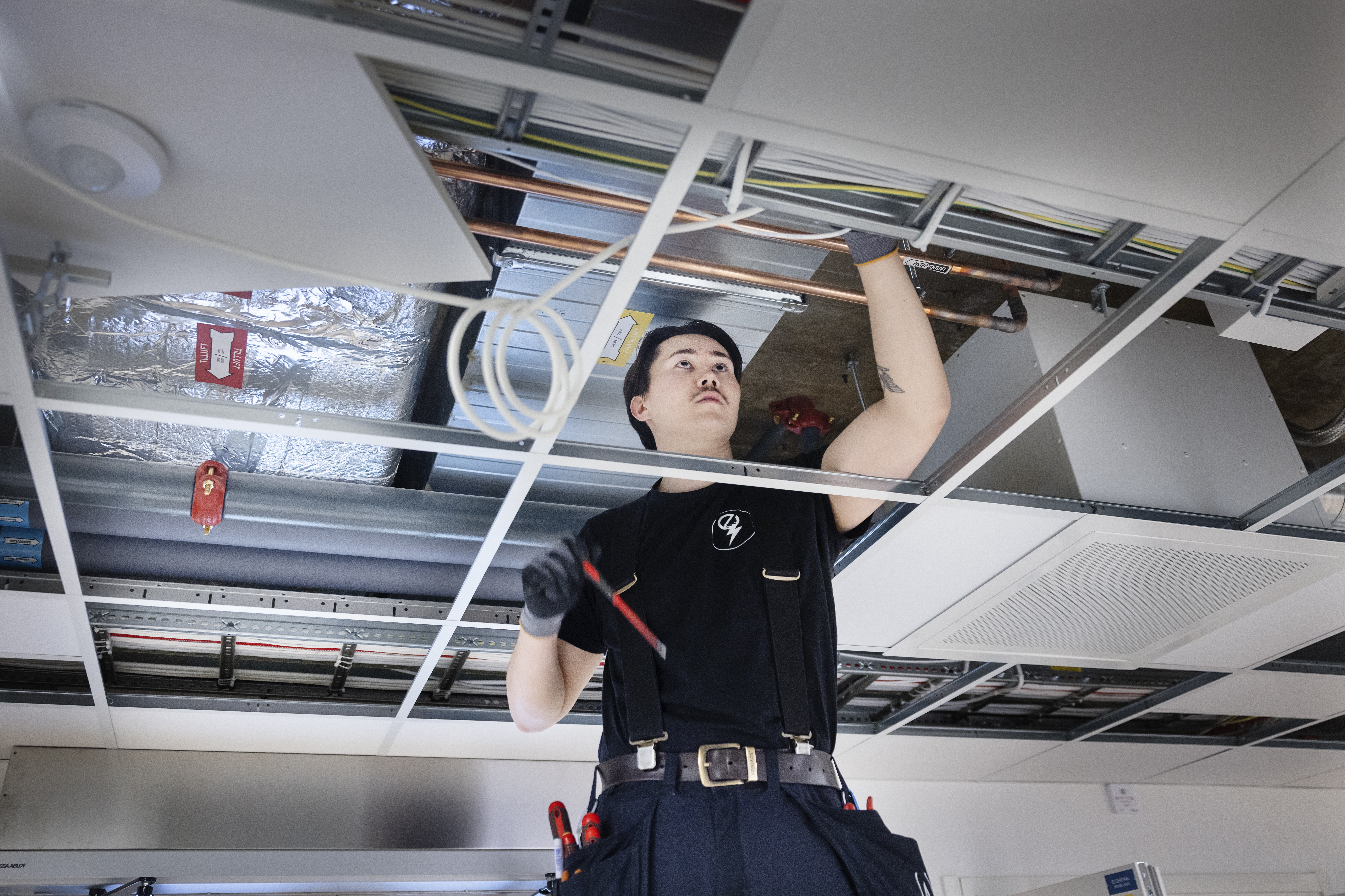 En elektriker i uniform arbetar med kablar ovanför ett undertak, håller en skruvmejsel och har ett verktygsbälte på sig.