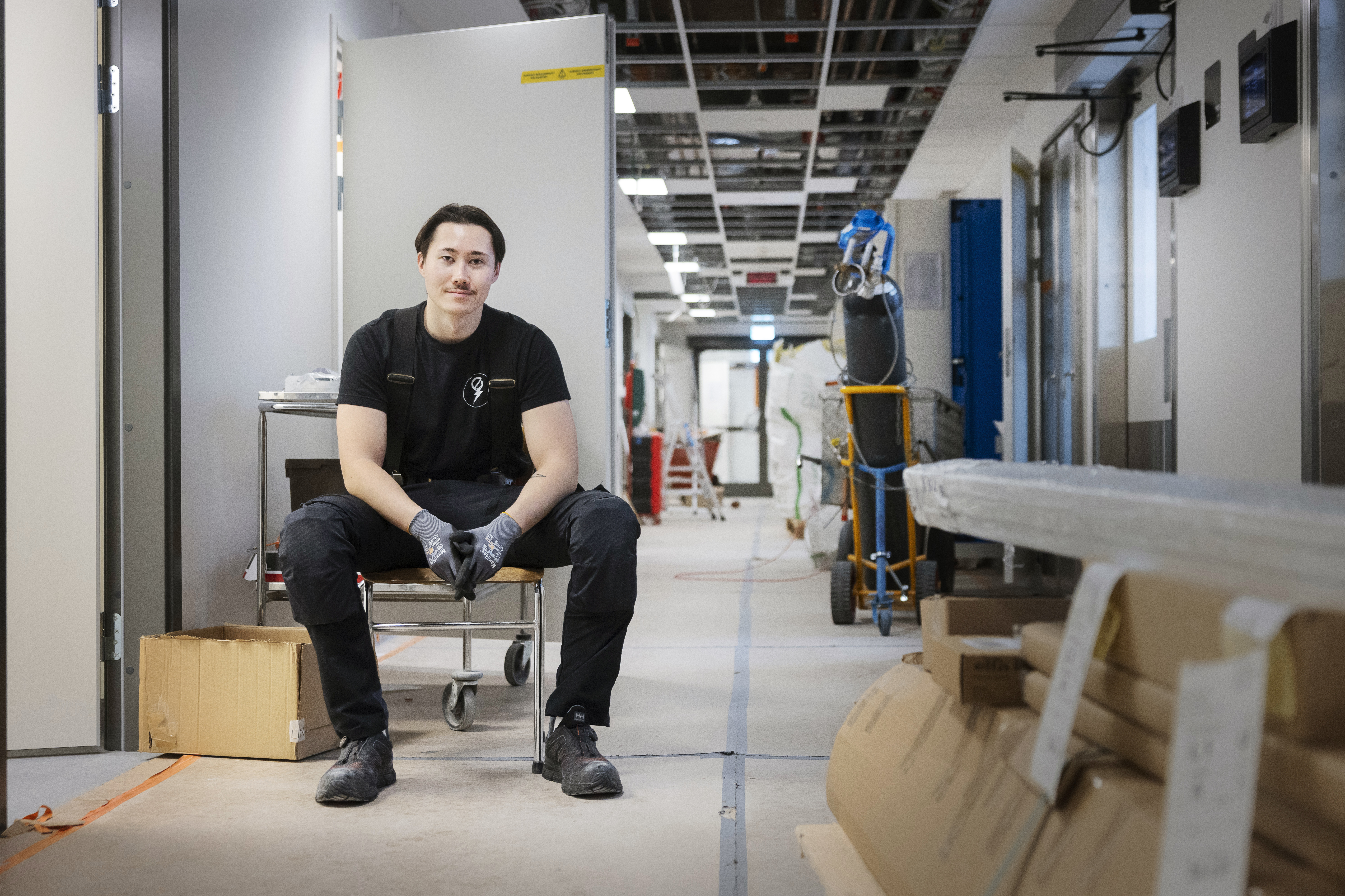 En person med arbetshandskar och svarta kläder sitter på en stol i en byggkorridor omgiven av lådor, verktyg och material.