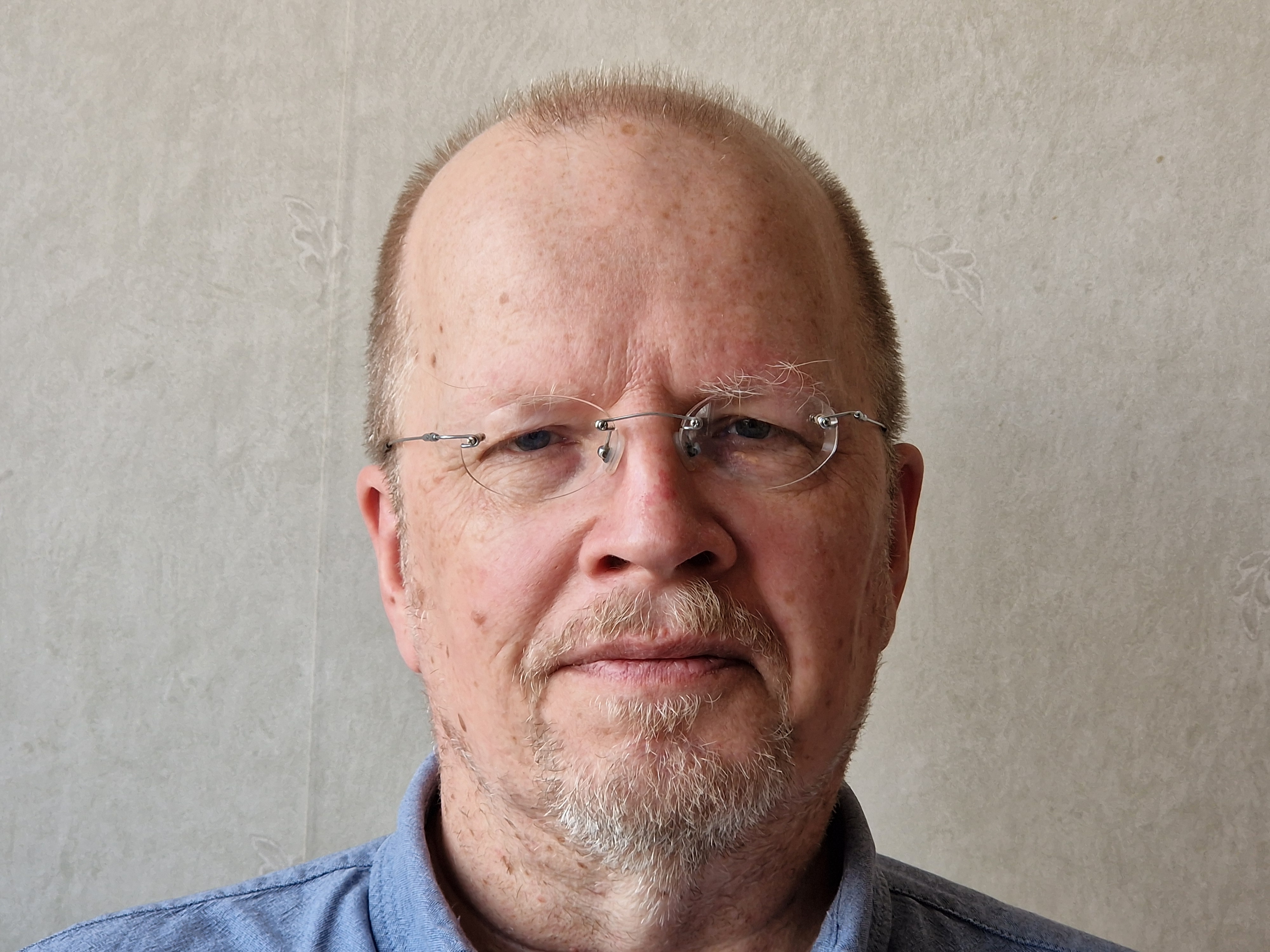 En man i medelåldern med glasögon, kort skägg och tunt hår står i blå skjorta framför en ljus vägg.