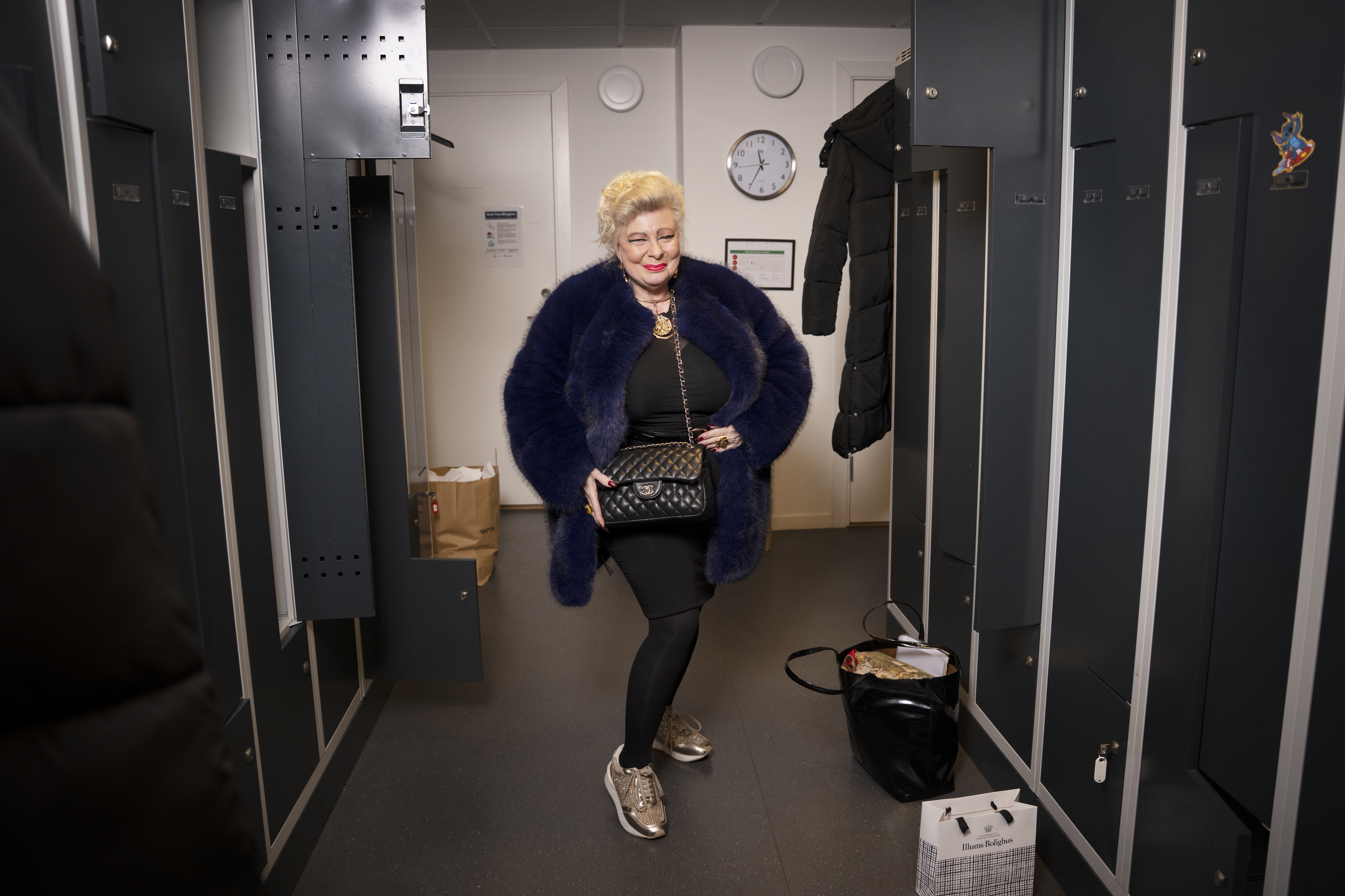 En äldre kvinna med en blå pälskappa, svarta kläder och sneakers står i ett omklädningsrum med skåp, väskor och klocka bakom sig.