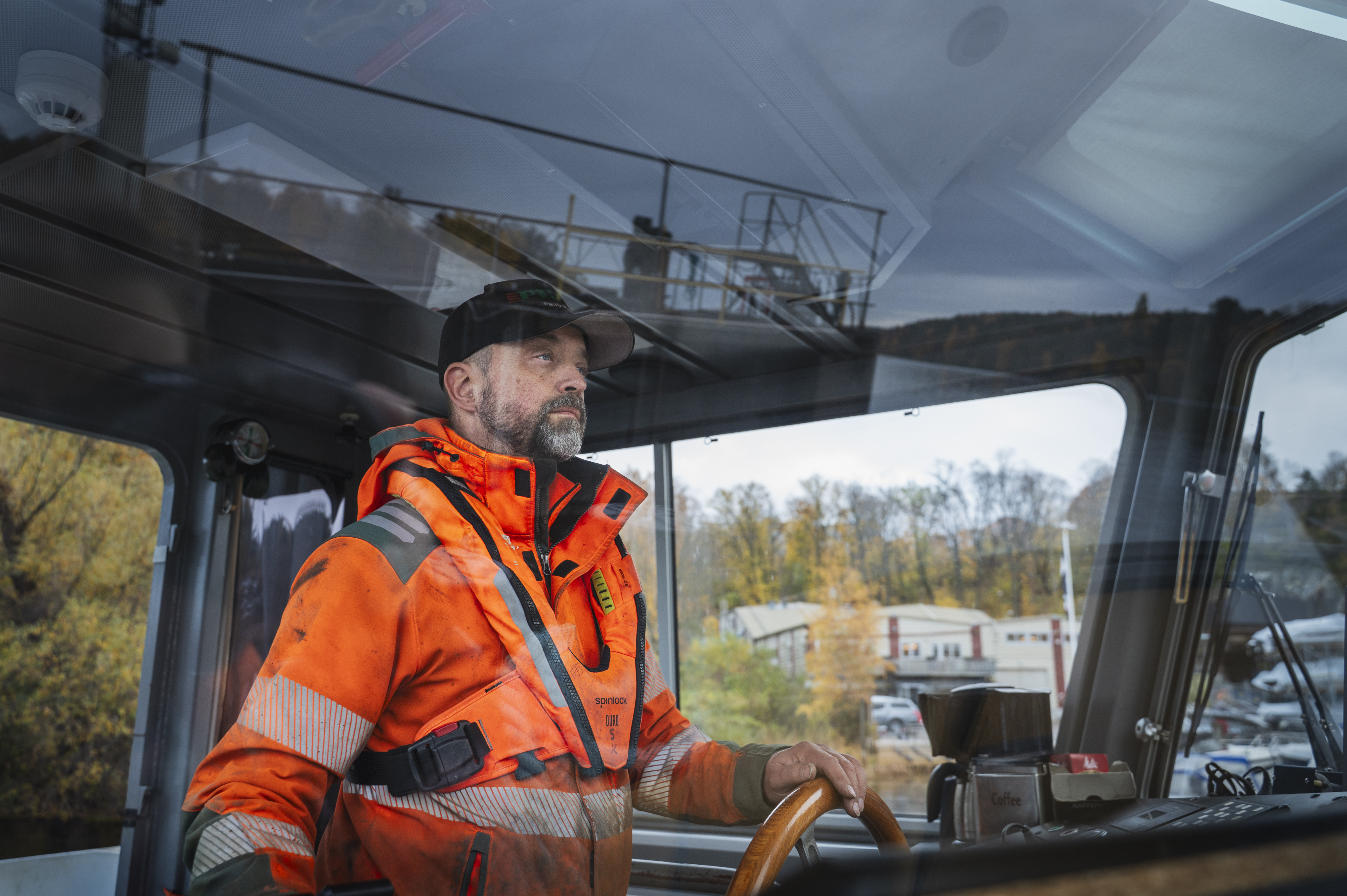 En person i orange säkerhetsjacka står vid rodret inne i en båt, styr och tittar framåt. Träd och byggnader syns genom fönstret.
